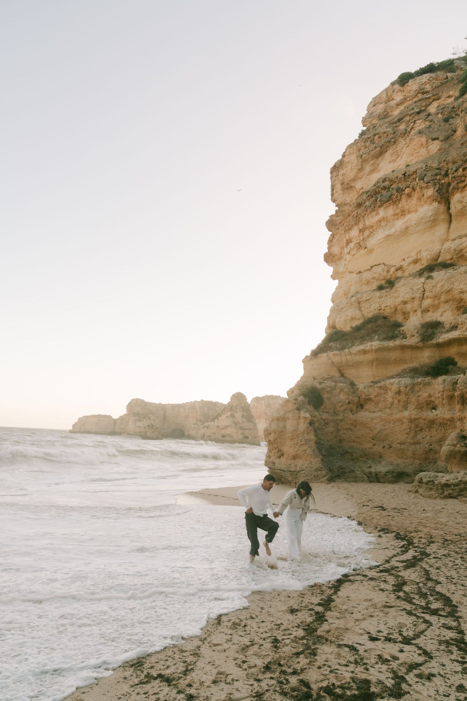 Couple playing in ocean waves on a sandy beach beside dramatic golden sandstone cliffs at dusk.