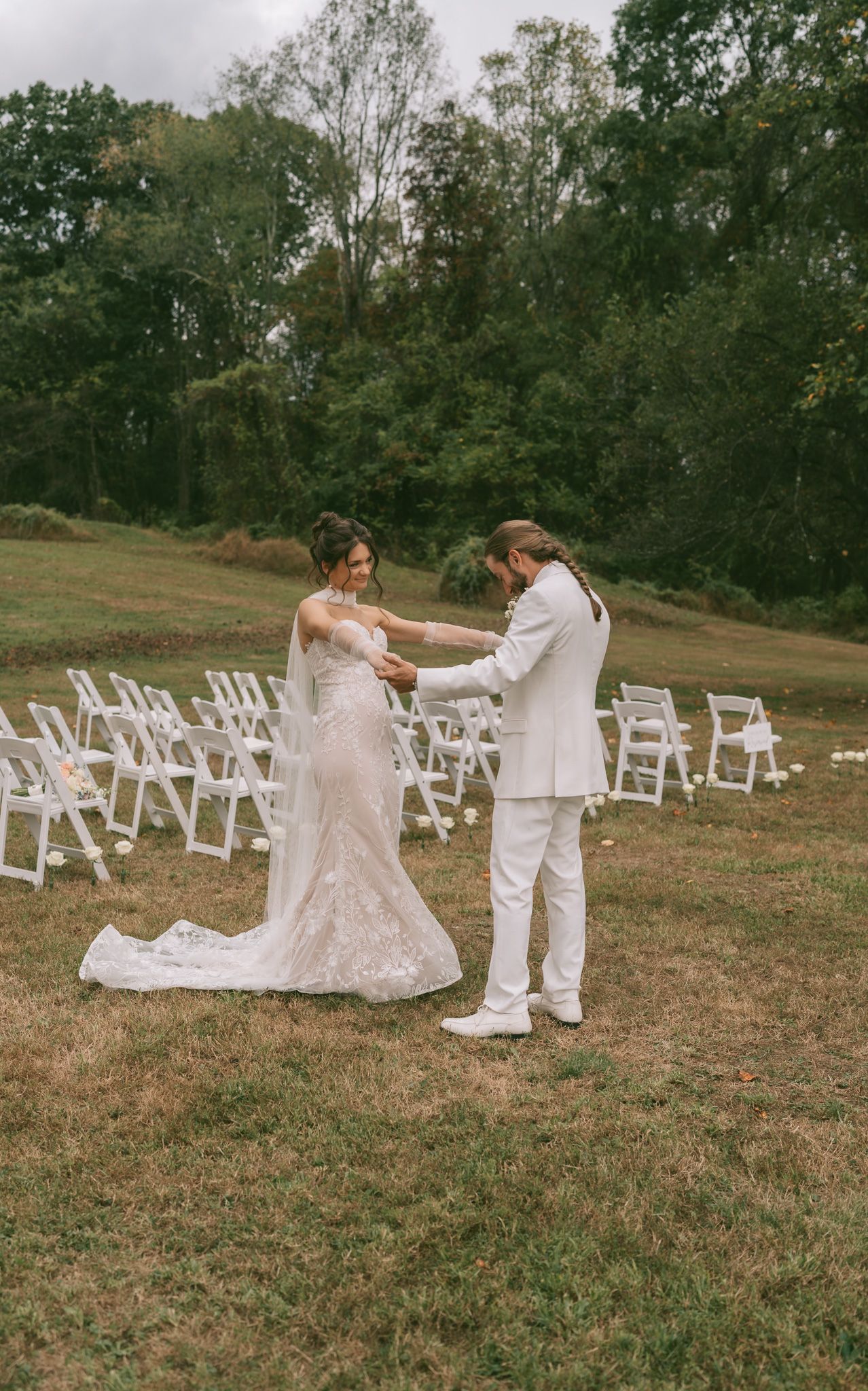 A couple in white attire dance together in a grassy field with empty white chairs arranged for a wedding ceremony.
