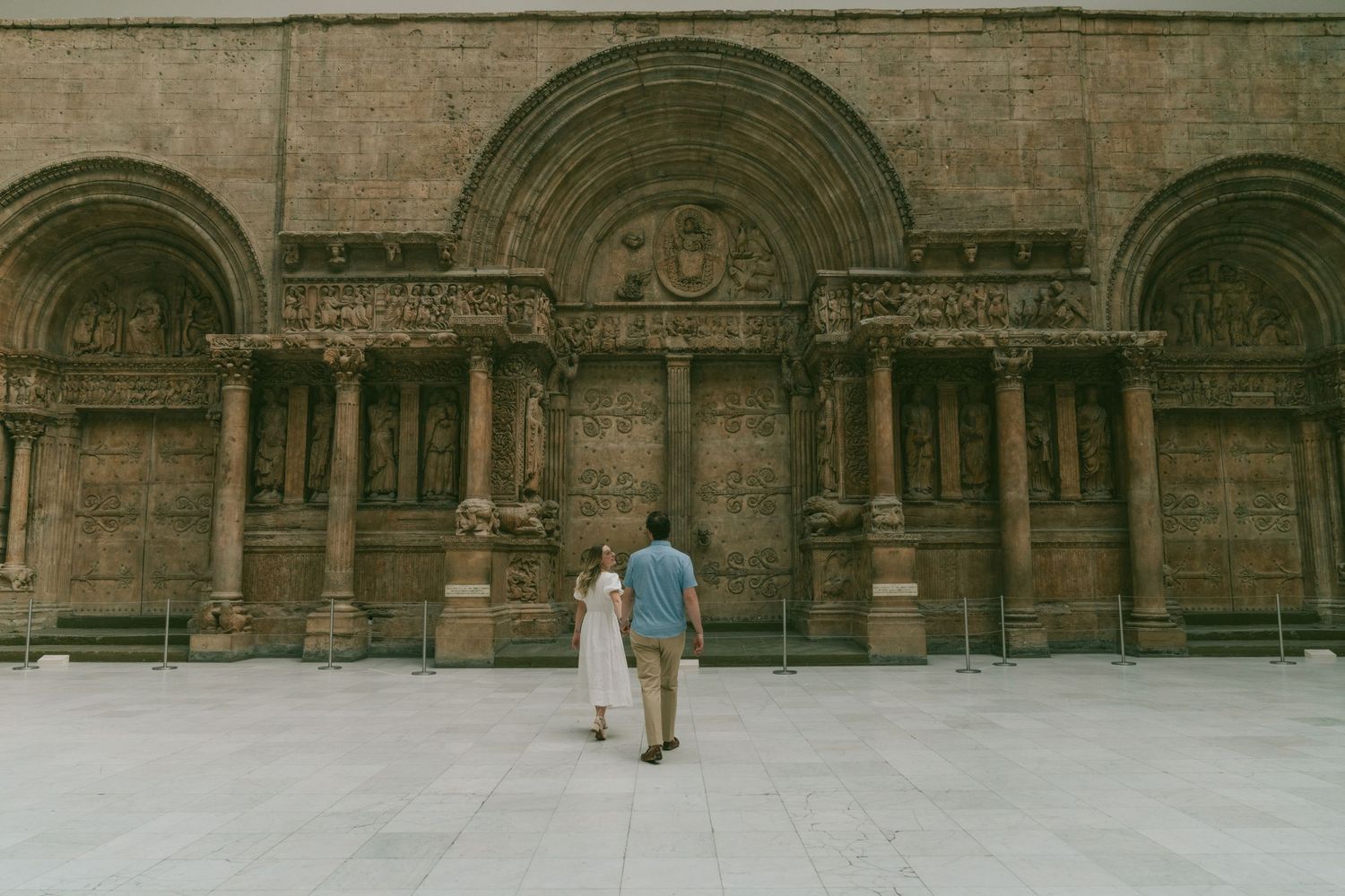 A couple stands before an ornate stone building facade with three arched entrances and classical architectural details.