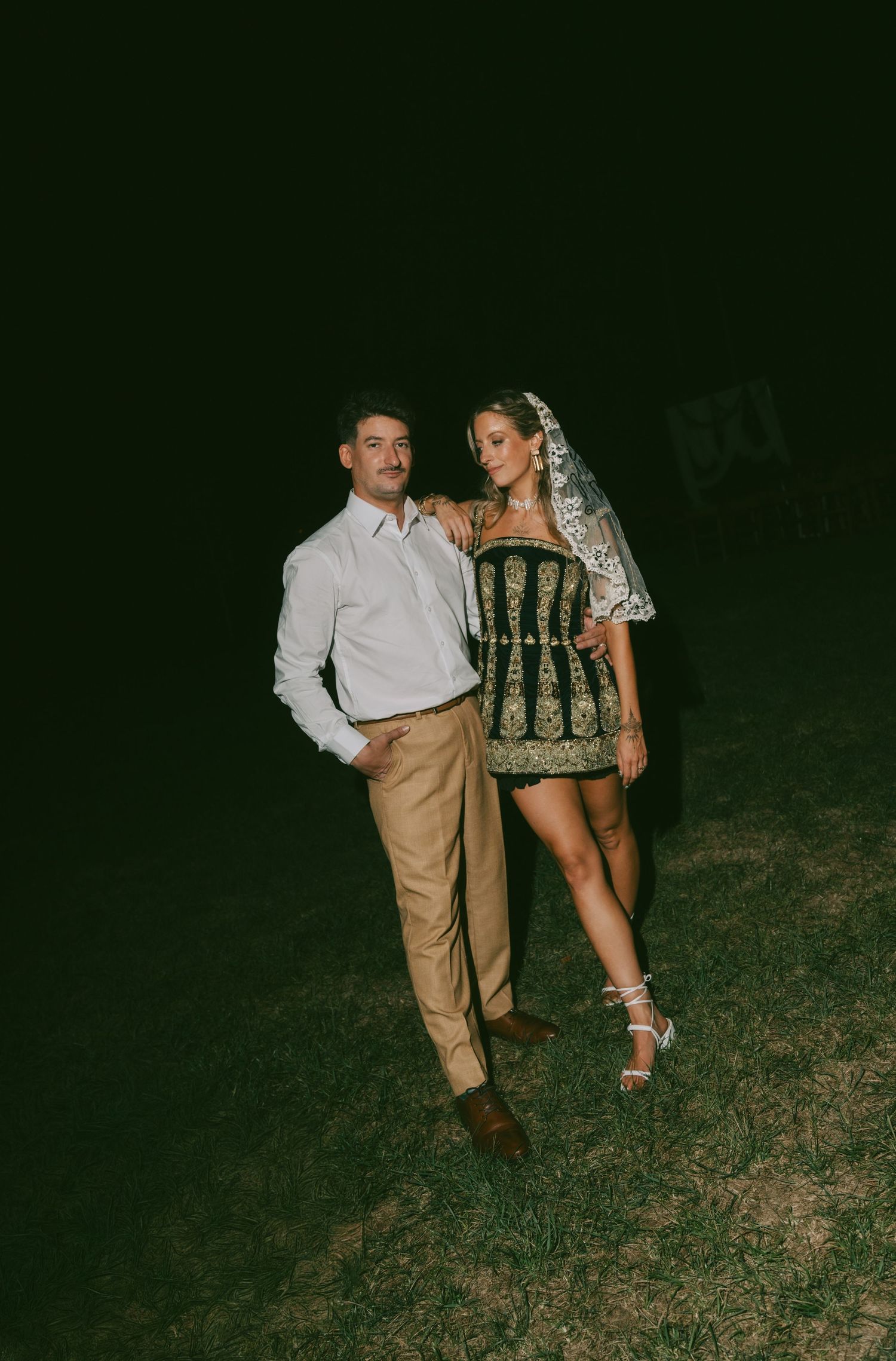 A couple poses together at night outdoors, with the woman wearing an ornate green and gold dress with a veil.