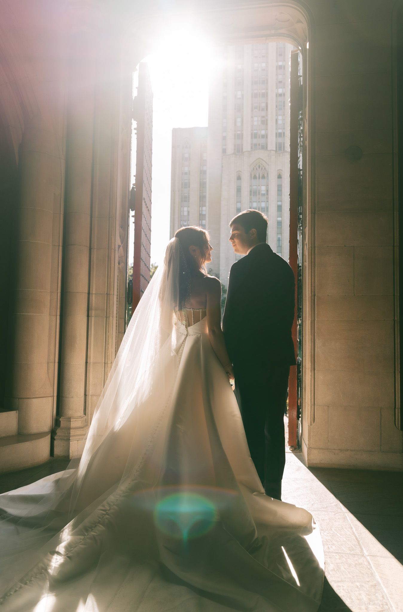 Silhouetted bride and groom stand in bright sunlit hallway with flowing veil creating ethereal wedding atmosphere.
