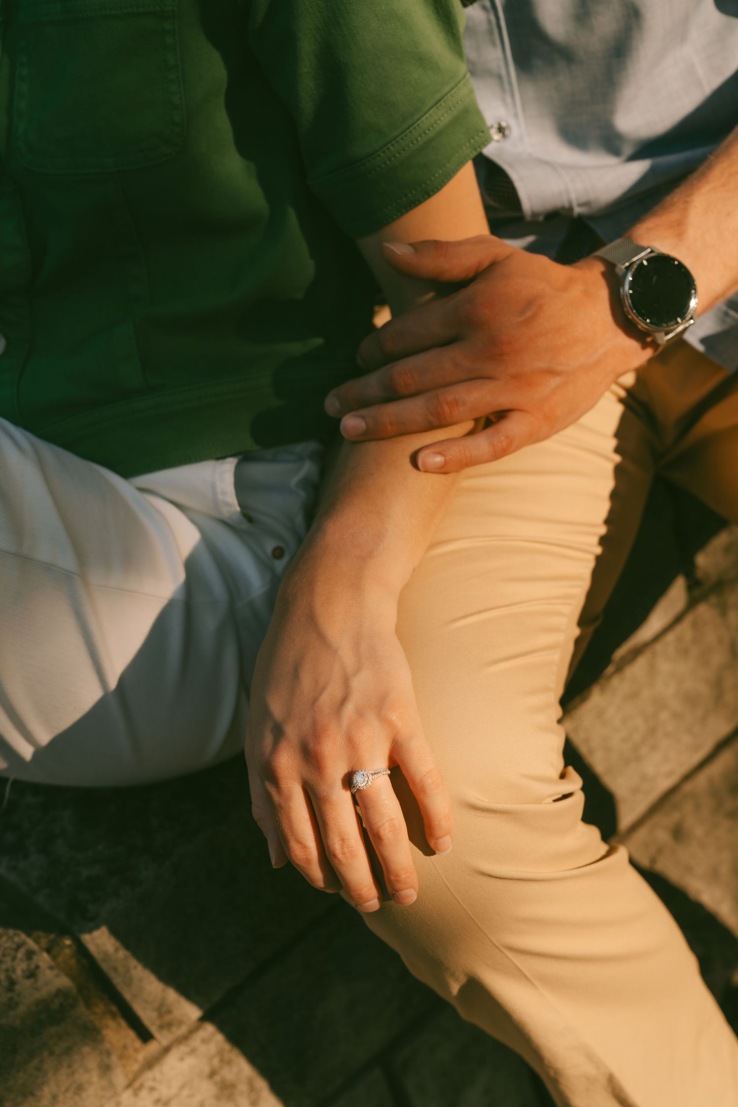 Close-up view of hands wearing a watch resting on beige pants with a green shirt visible in natural lighting.