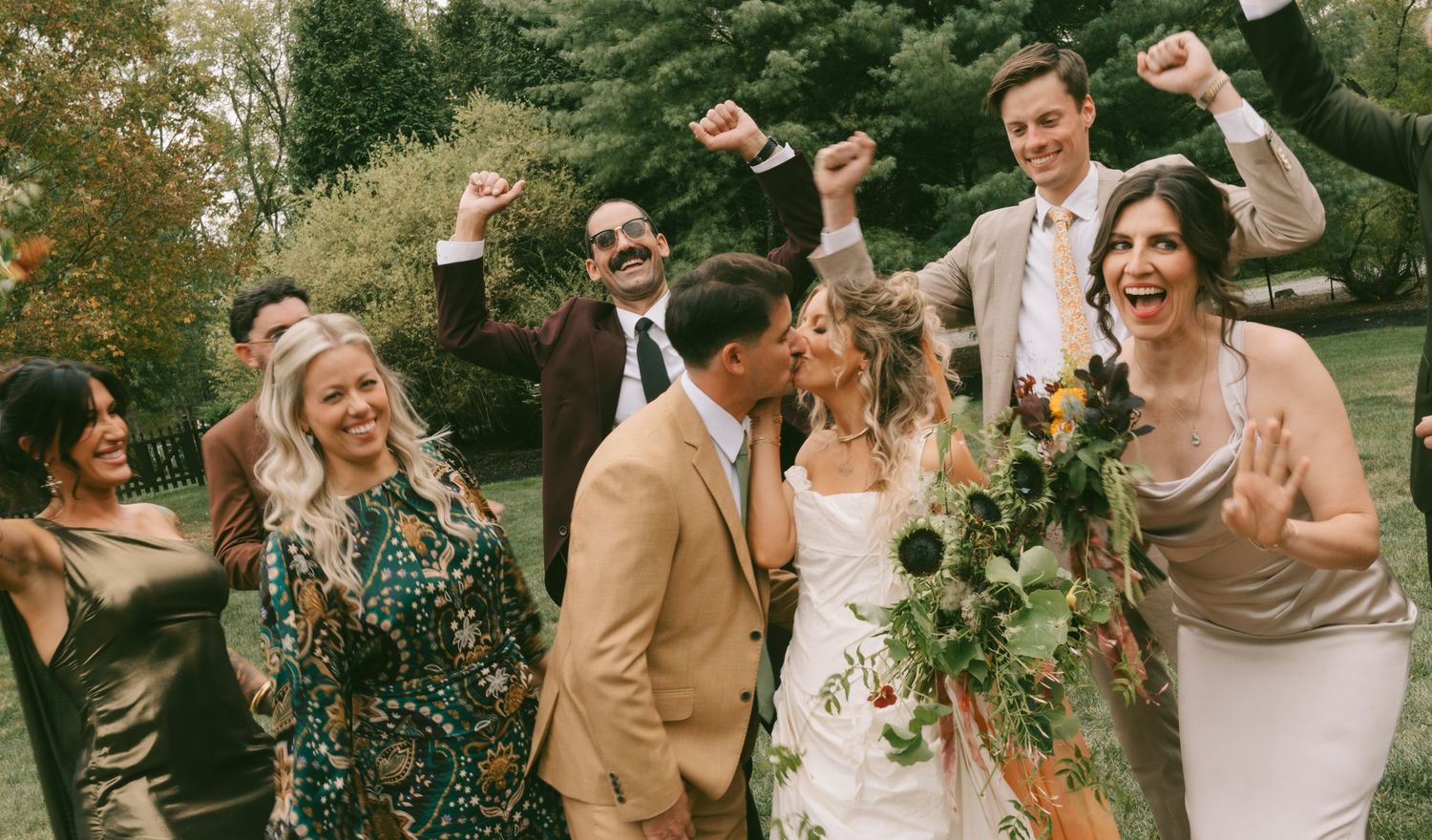 Wedding guests celebrate joyfully in a garden setting with animated expressions and raised arms.