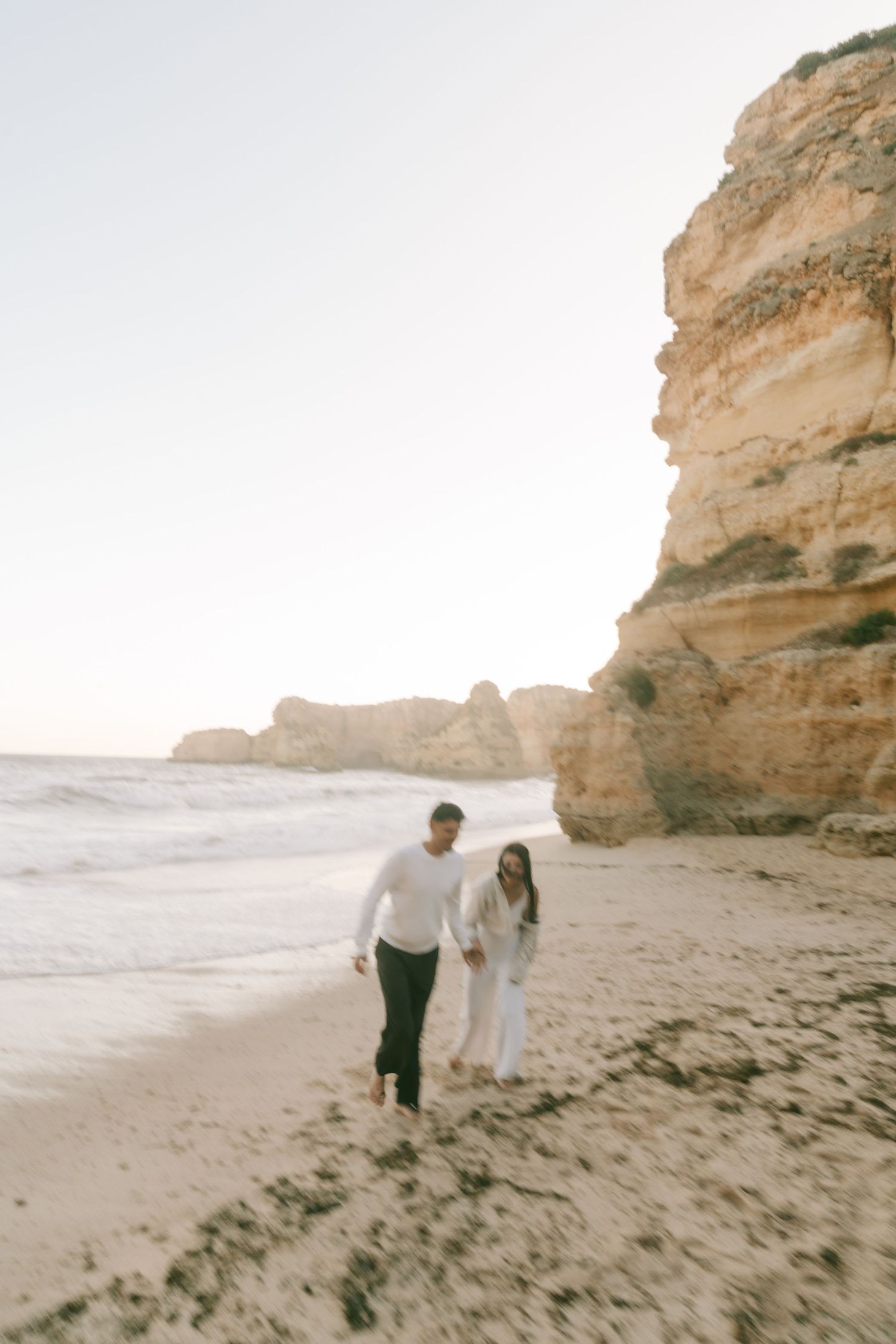 Couple walking hand in hand along a sandy beach beside tall golden sandstone cliffs at sunset.