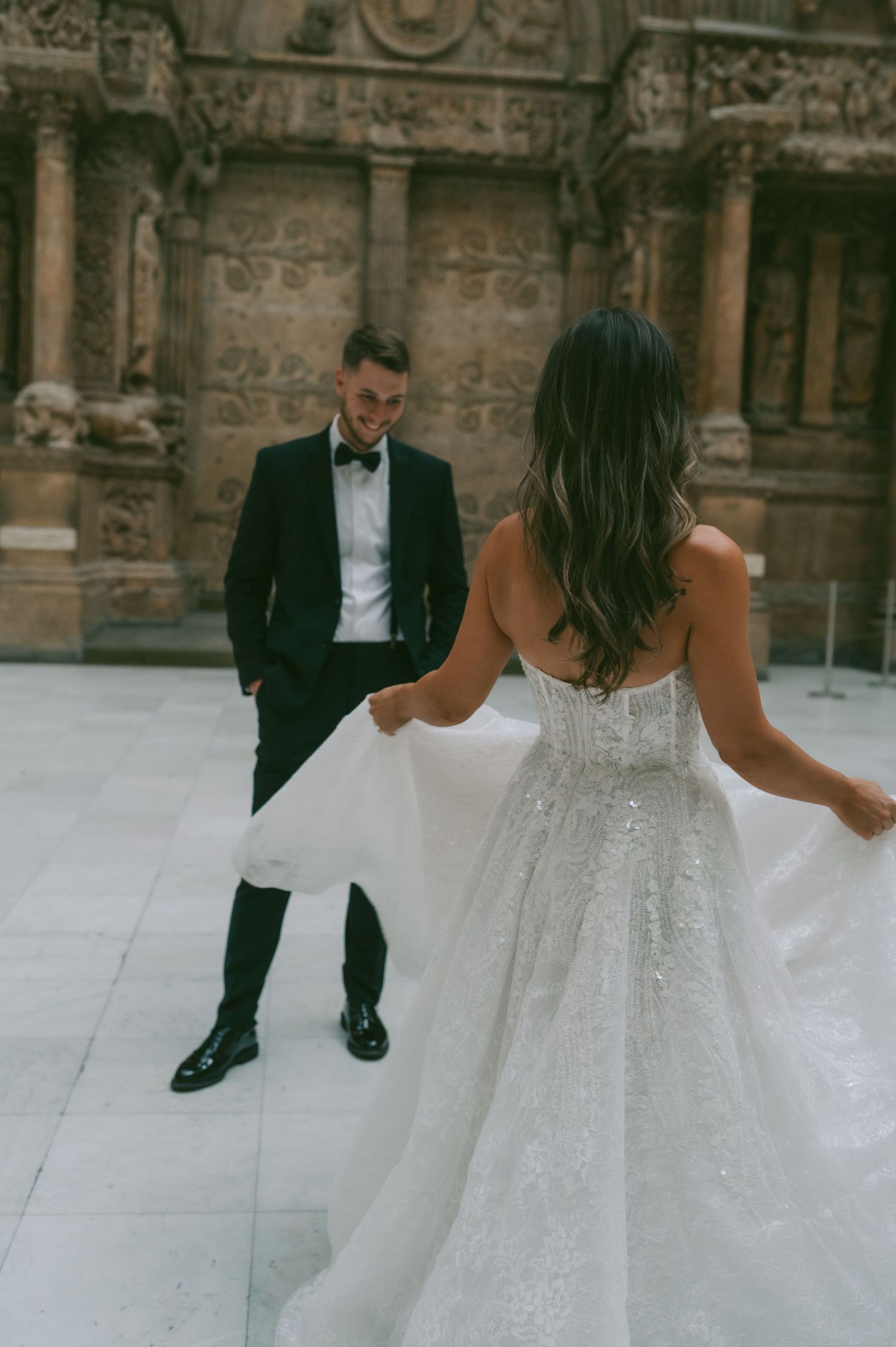 A bride in a flowing white dress and groom in black tuxedo pose together in front of an ornate stone building.