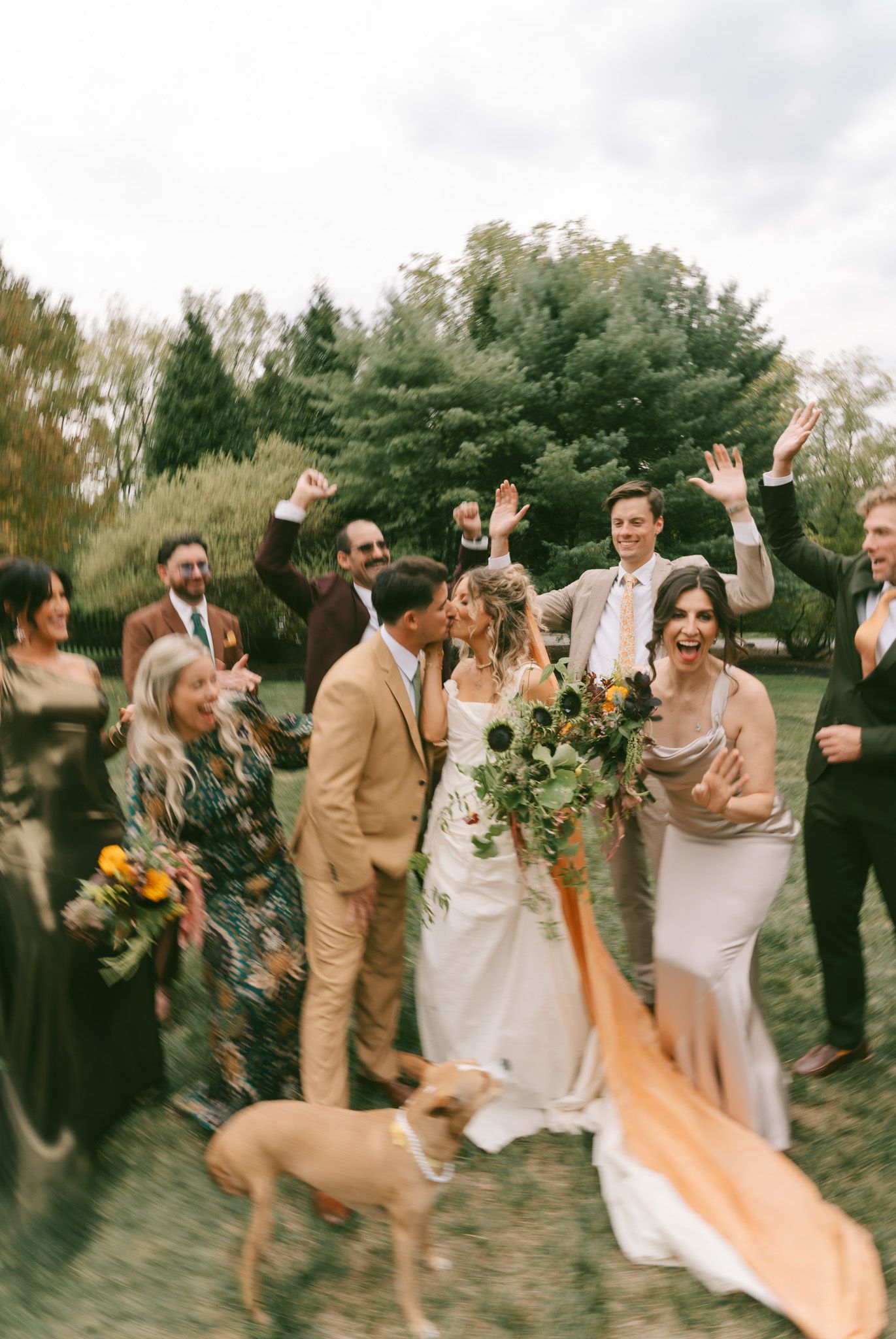 Wedding party celebrates outdoors with joyful gestures while a dog walks through the festive scene.