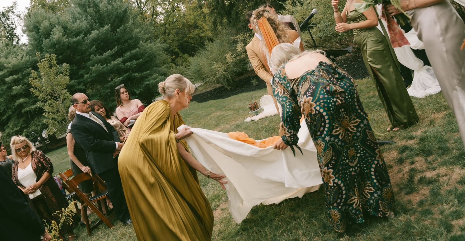 Wedding guests in vintage-style dresses help carry a flowing white dress train across a grassy outdoor setting.