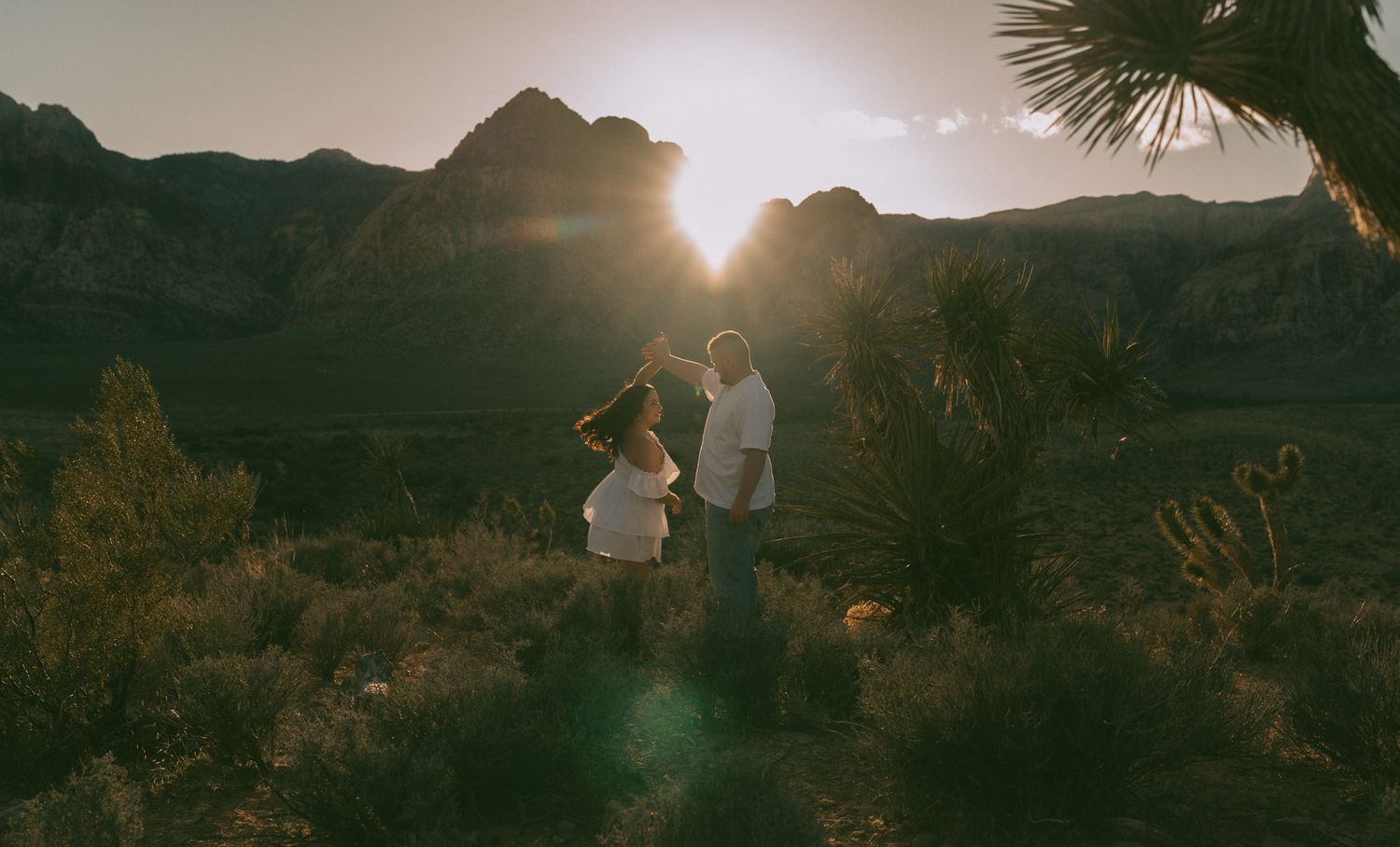 Two figures stand against a silhouetted mountainous backdrop during golden hour sunset.