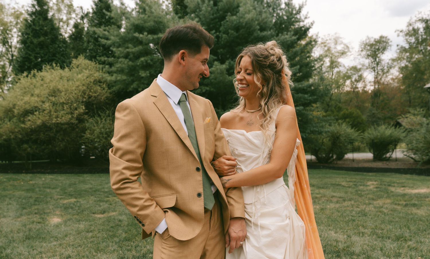 A couple shares a joyful moment together outdoors in wedding attire, with the bride in white and groom in tan.