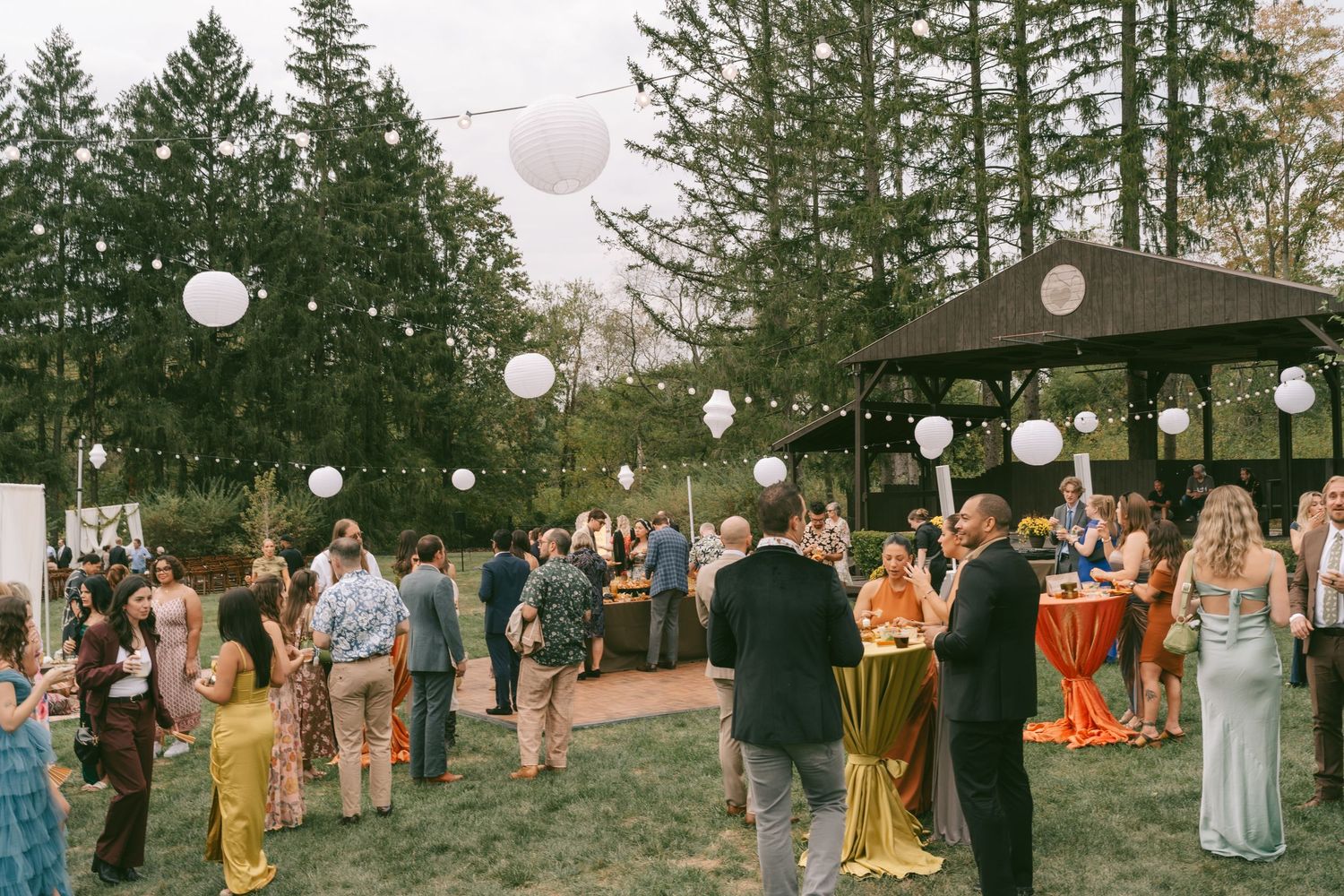 Wedding guests gather outdoors under white paper lanterns with evergreen trees in the background at dusk.