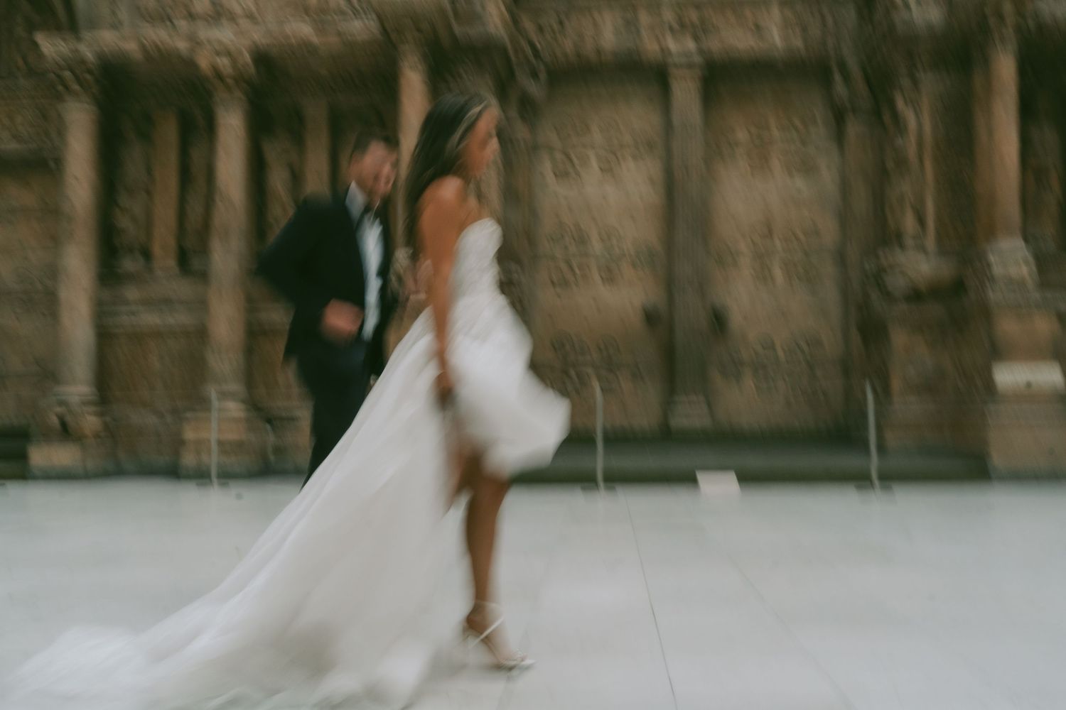 Newlywed couple dances gracefully across a grand European courtyard with historic architecture in the background.