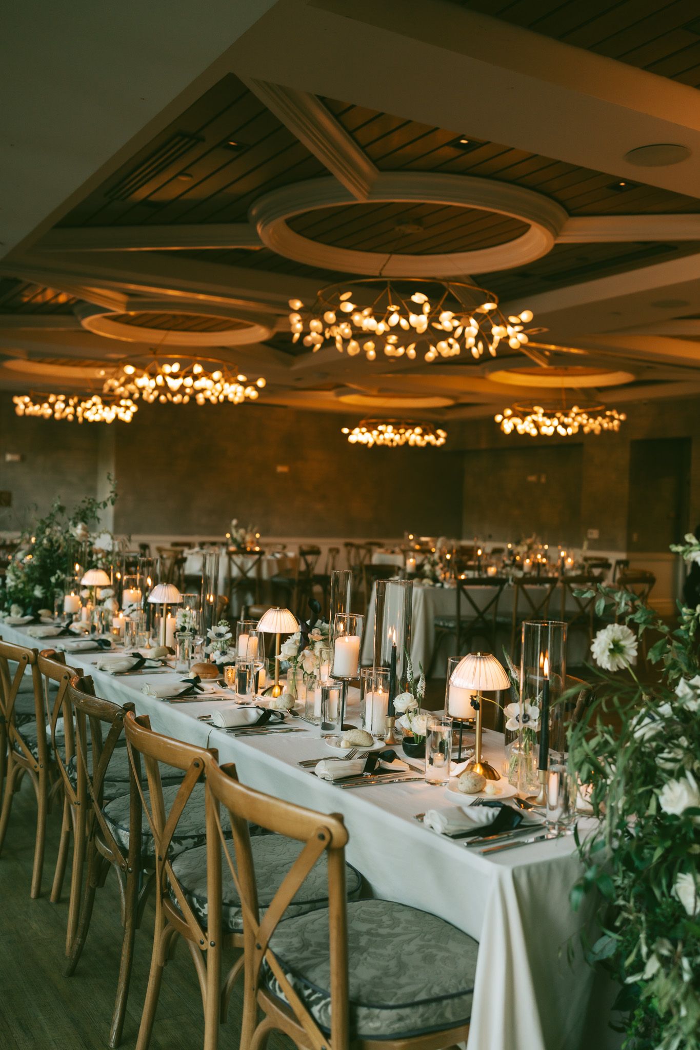 Elegant wedding reception table with white linens, wooden chairs, and ornate chandeliers casting warm light from above.