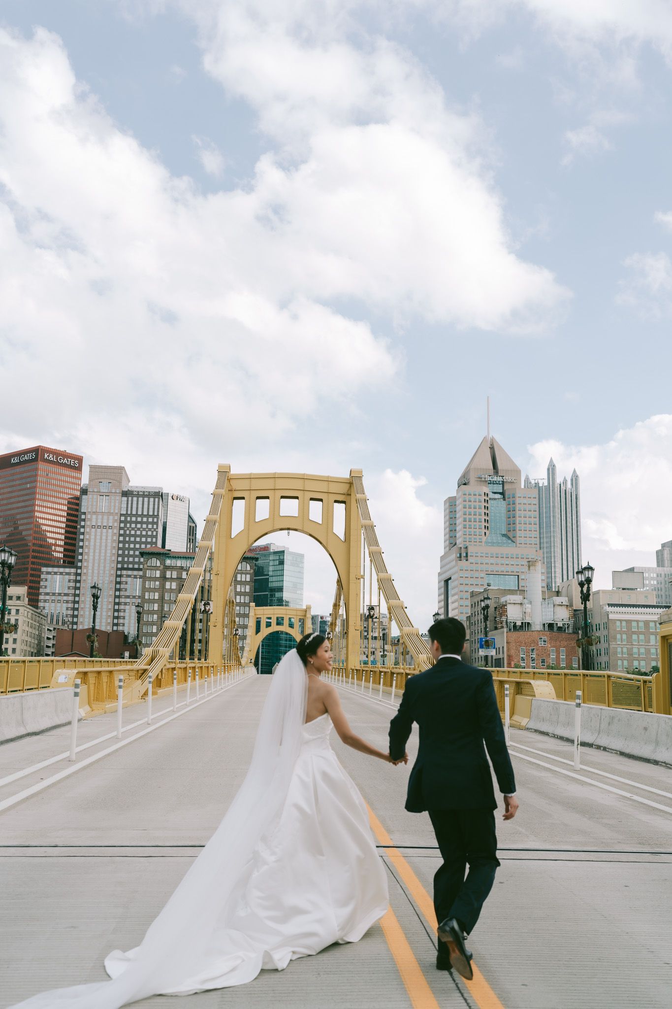 Bride and groom walk along Pittsburgh's iconic yellow Roberto Clemente Bridge with city skyline in background.