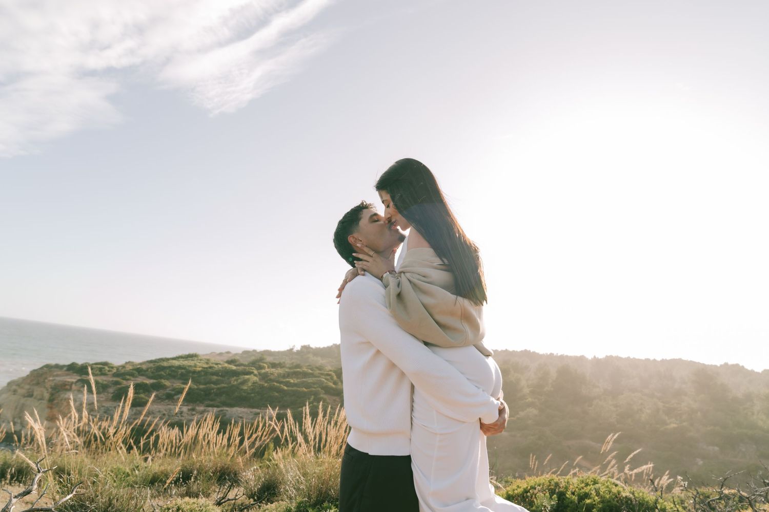 Romantic couple embracing and kissing on a scenic coastal hillside at golden hour sunset.