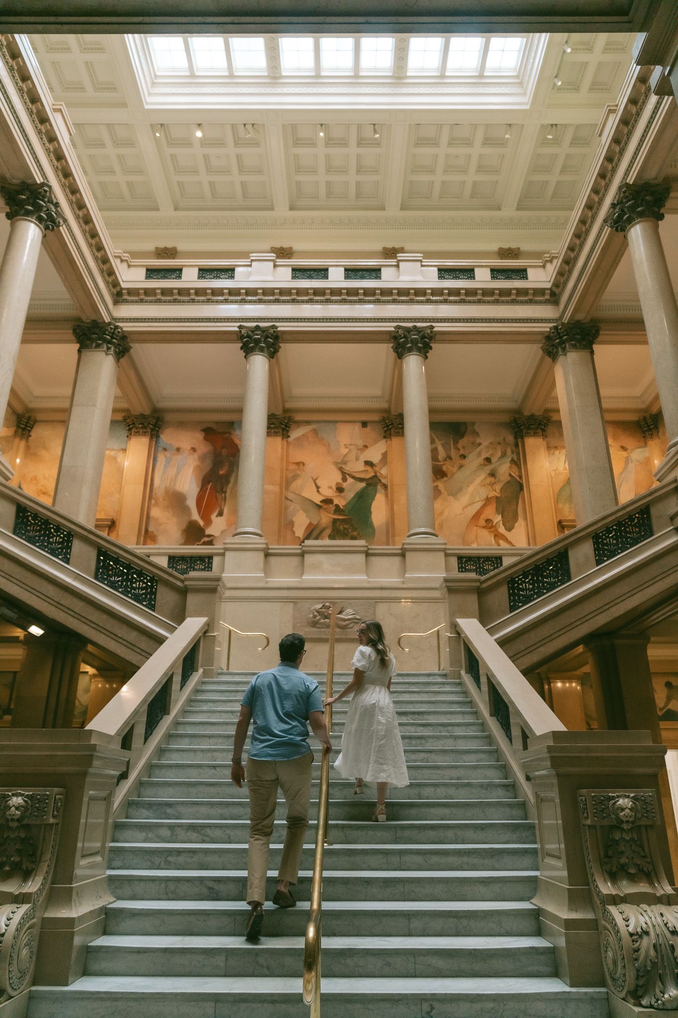 Couple ascending grand marble staircase in museum with classical columns and skylight.