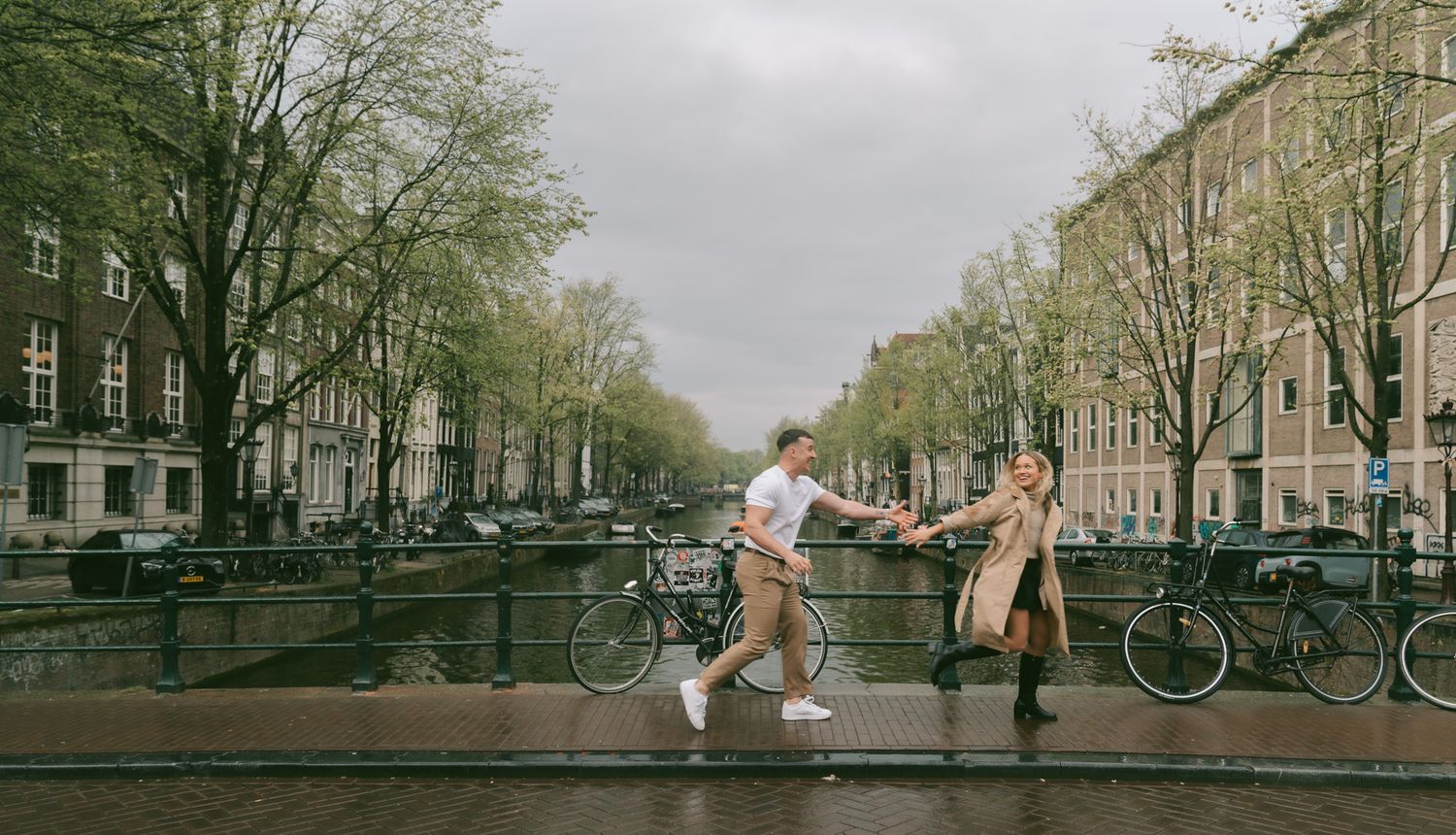 Playful couple jumping on Amsterdam canal bridge surrounded by autumn trees.