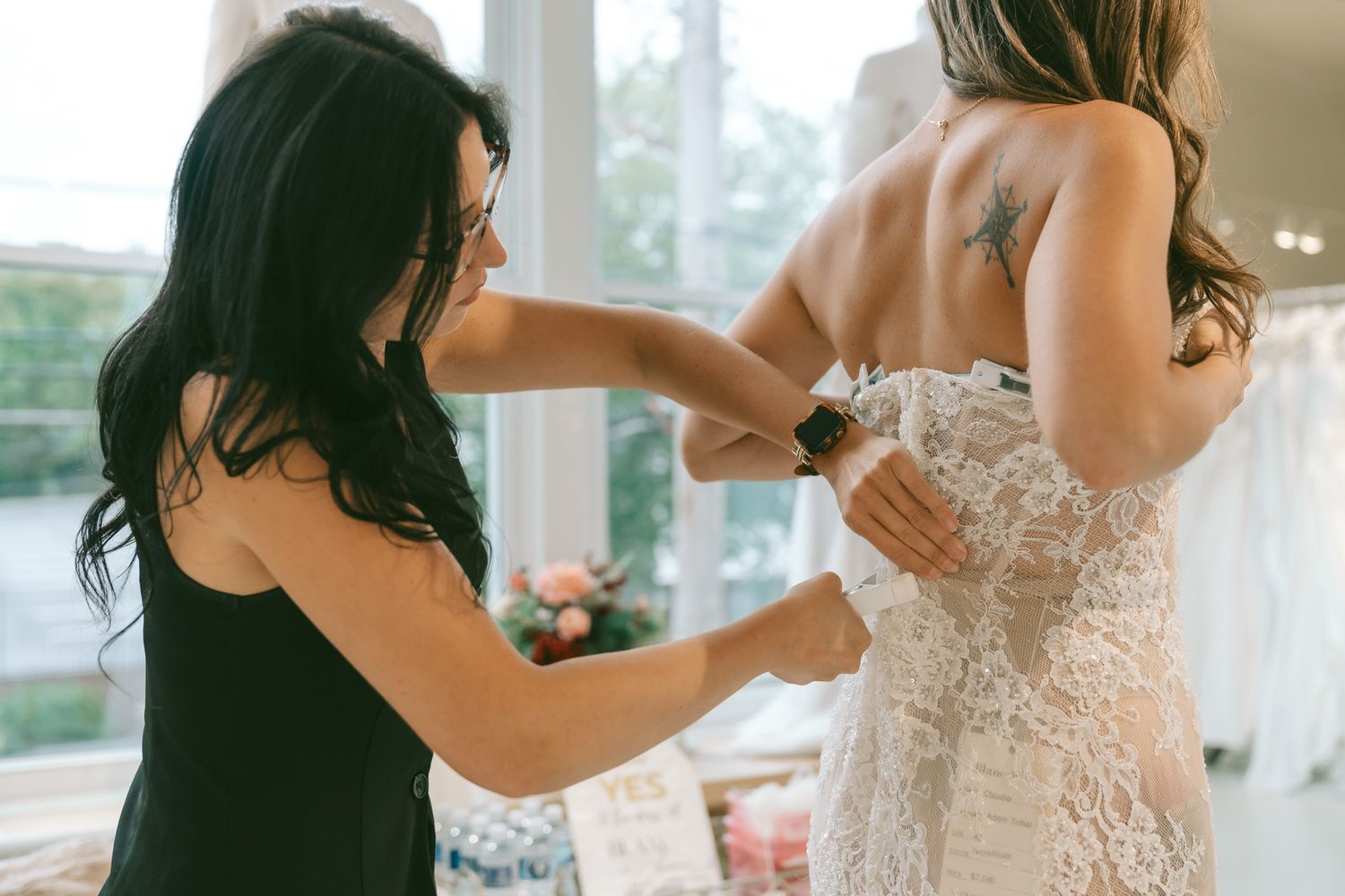 A bride gets help putting on her elegant white lace wedding dress in a sunlit room.