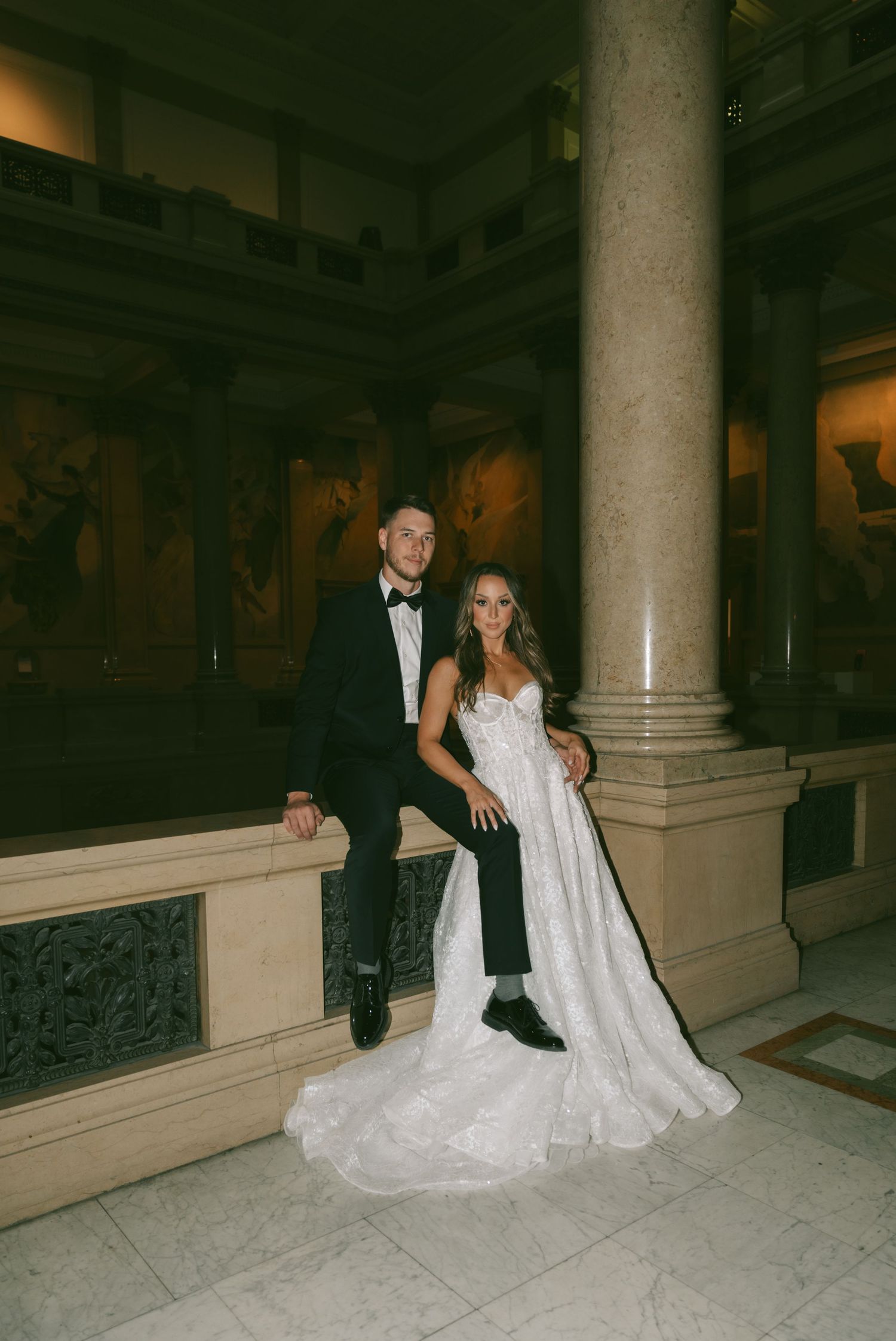 Couple in formal wedding attire posing on marble staircase in elegant venue with classical architecture.