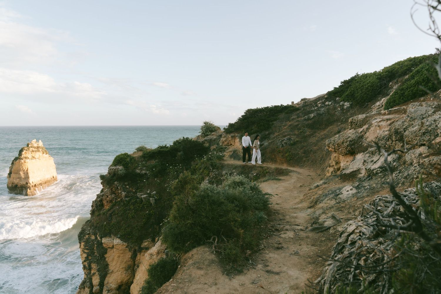 Couple walking on a scenic coastal cliff path overlooking rocky sea stacks and ocean waves at golden hour.