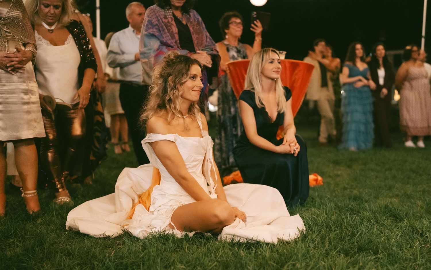 Wedding guests gathered on a lawn during sunset with festive string lights overhead.