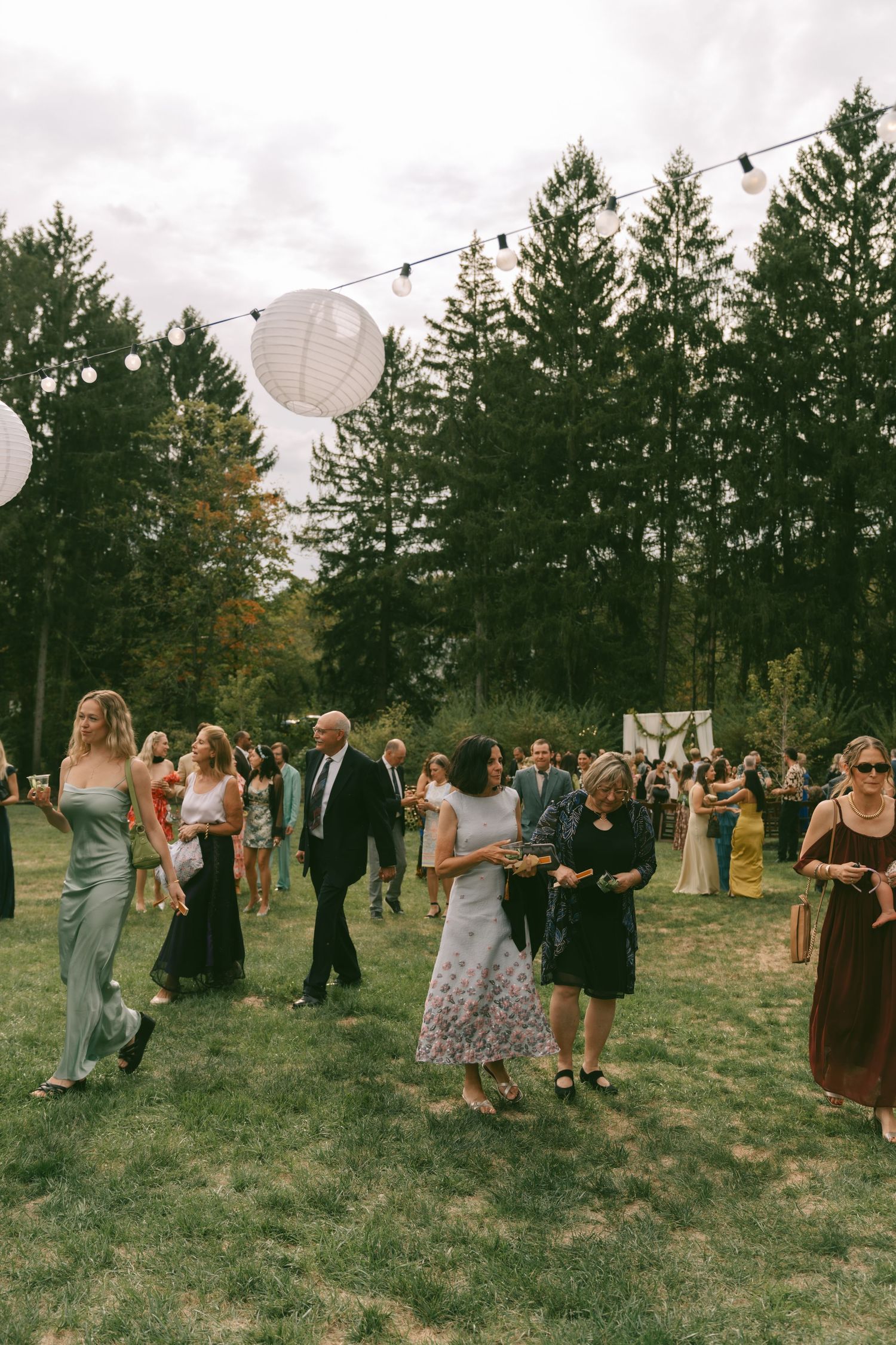 Guests mingle and dance at an outdoor wedding reception with string lights and paper lanterns among evergreen trees.