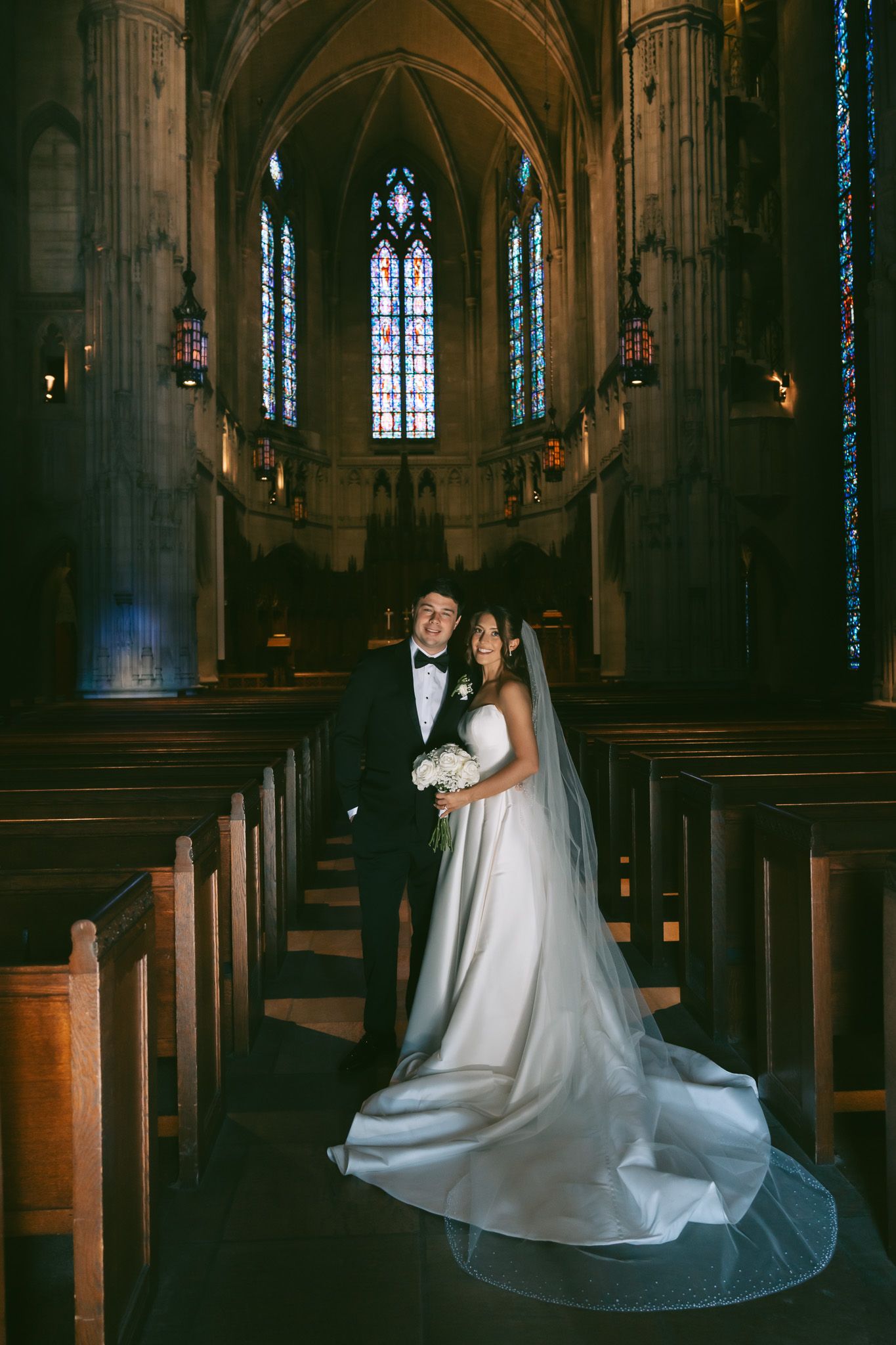 A romantic wedding couple poses in a grand gothic cathedral with dramatic lighting and stained glass windows in the background.