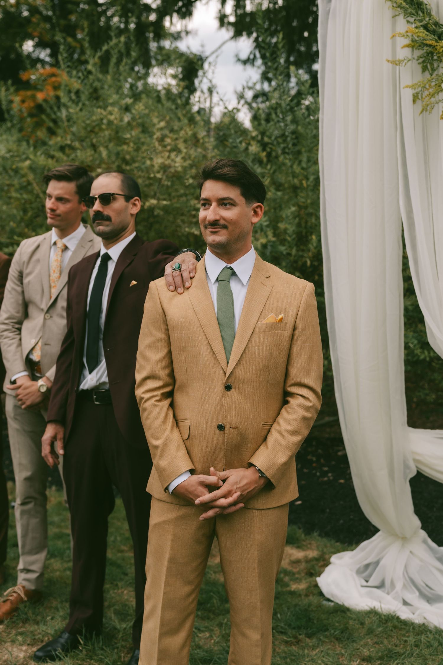 Groomsmen in tan and beige suits stand together under white draping at an outdoor wedding ceremony.