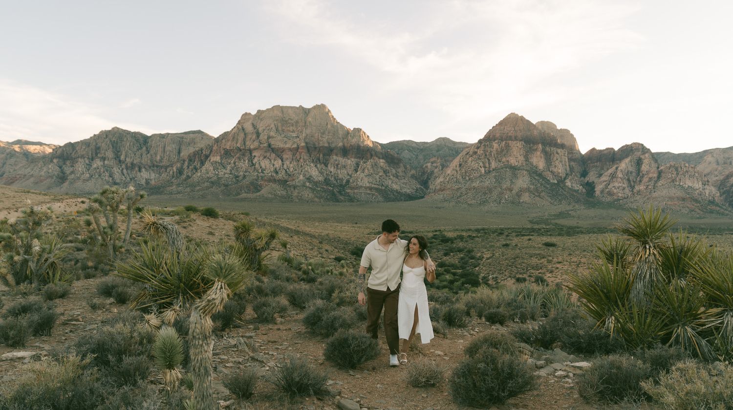 A couple stands together in the desert landscape of Red Rock Canyon, las vegas,  with mountains and Joshua trees in the background.