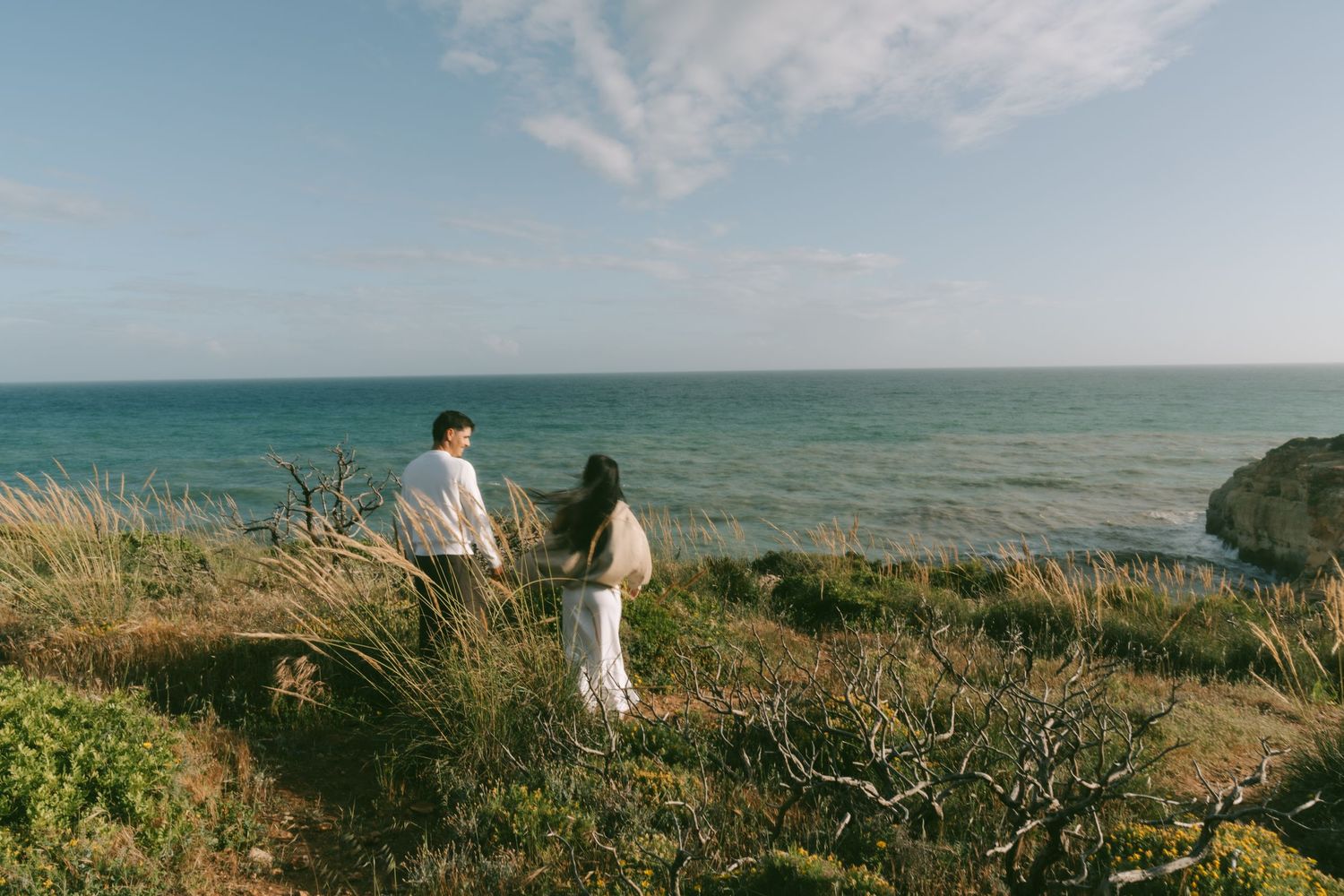 Couple standing on a coastal cliff overlooking the turquoise ocean, surrounded by wild grass and greenery.