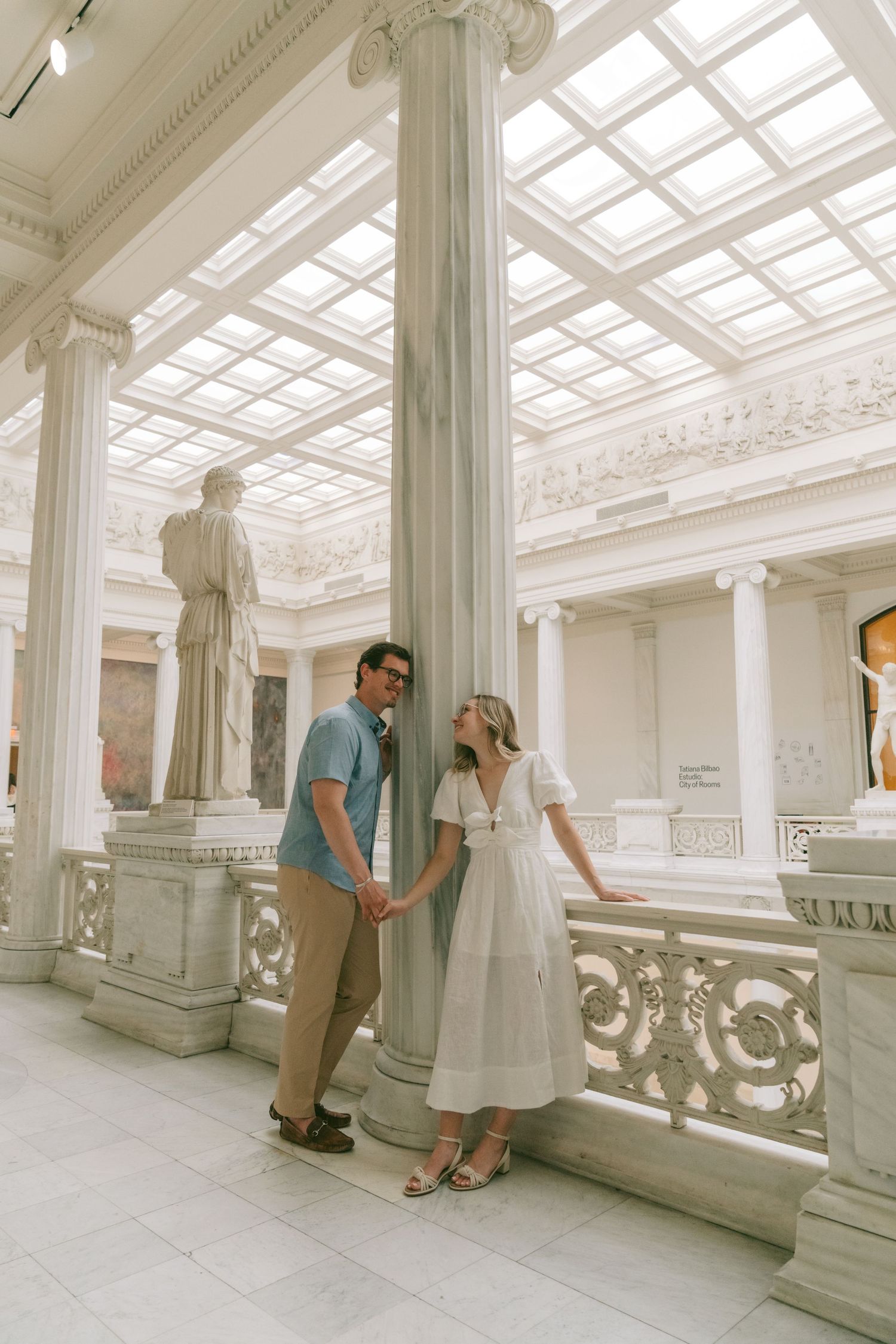 A couple shares a tender moment in an elegant white interior hallway with ornate architectural details and skylights.