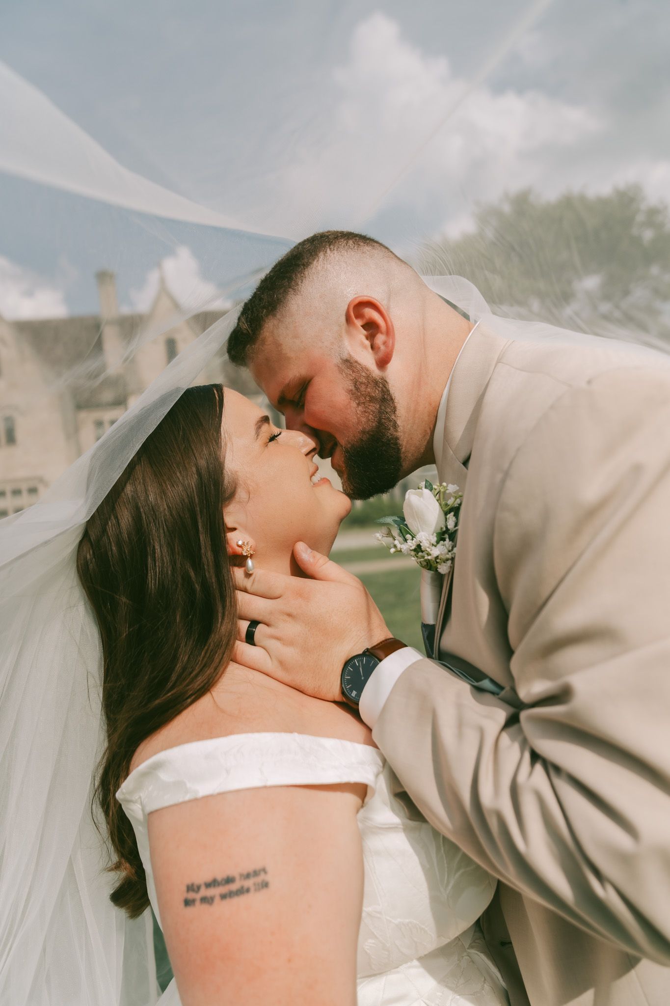 A romantic wedding couple shares a tender kiss under a flowing white veil outdoors.