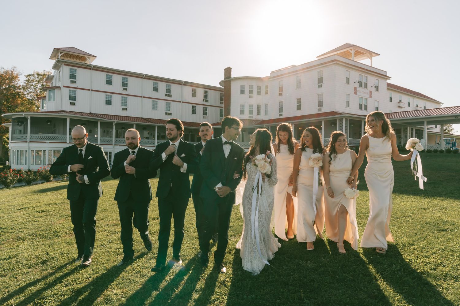 Wedding party in formal attire poses together on a sunny lawn in front of a grand white hotel building.