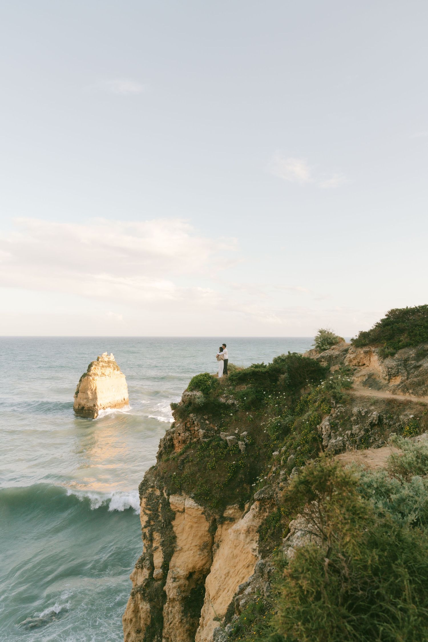 Couple standing on a dramatic coastal cliff overlooking turquoise ocean waves and a sea stack in the Algarve, Portugal.