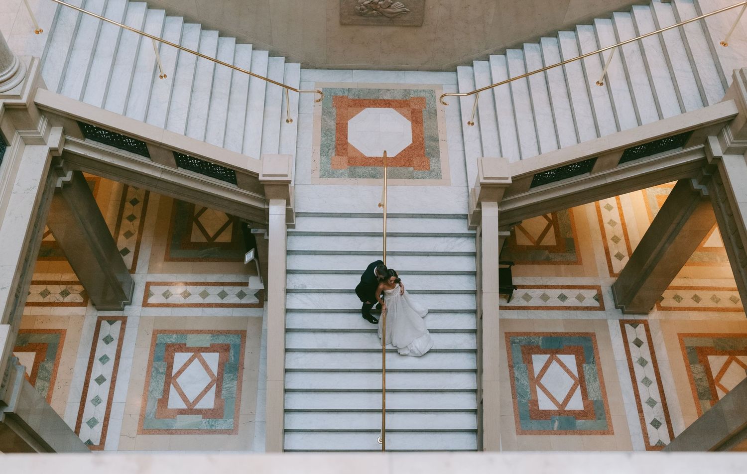 Looking down at a geometric staircase with ornate patterned floor tiles and a figure descending the white stairs.