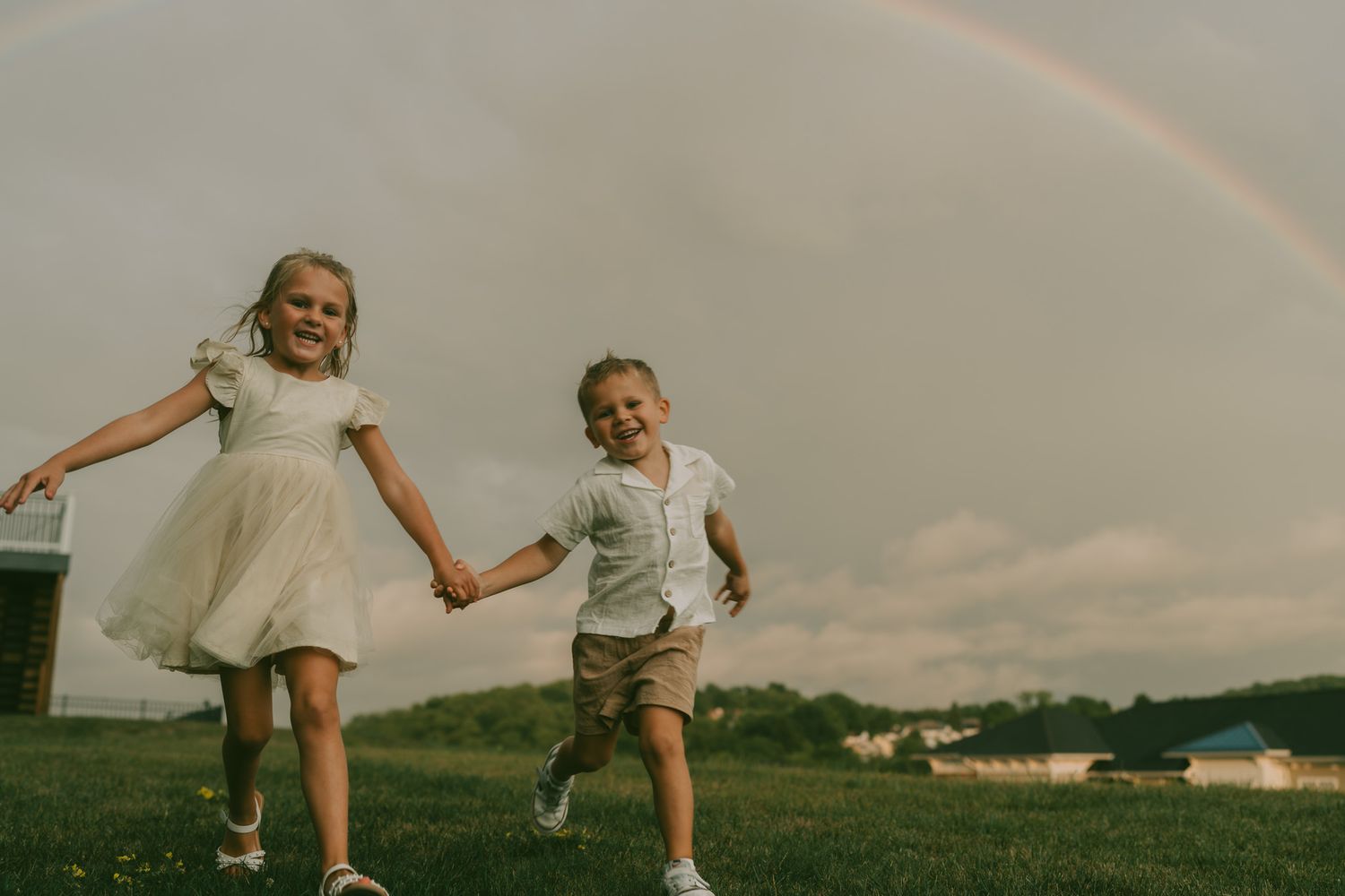 Children jumping playfully in a grassy field under a rainbow in the sky.