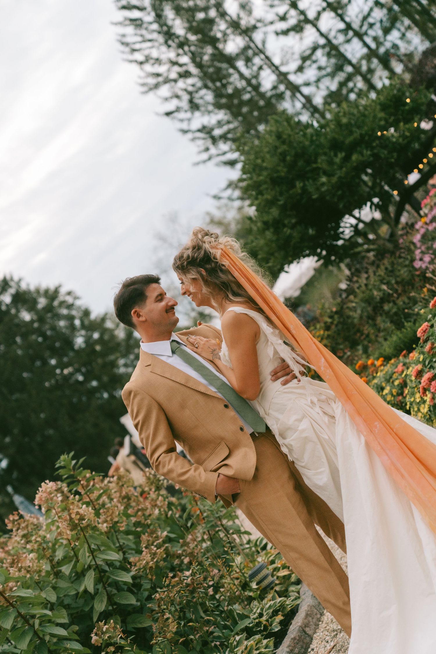 Romantic wedding couple embraces in garden setting with orange flowers and greenery.