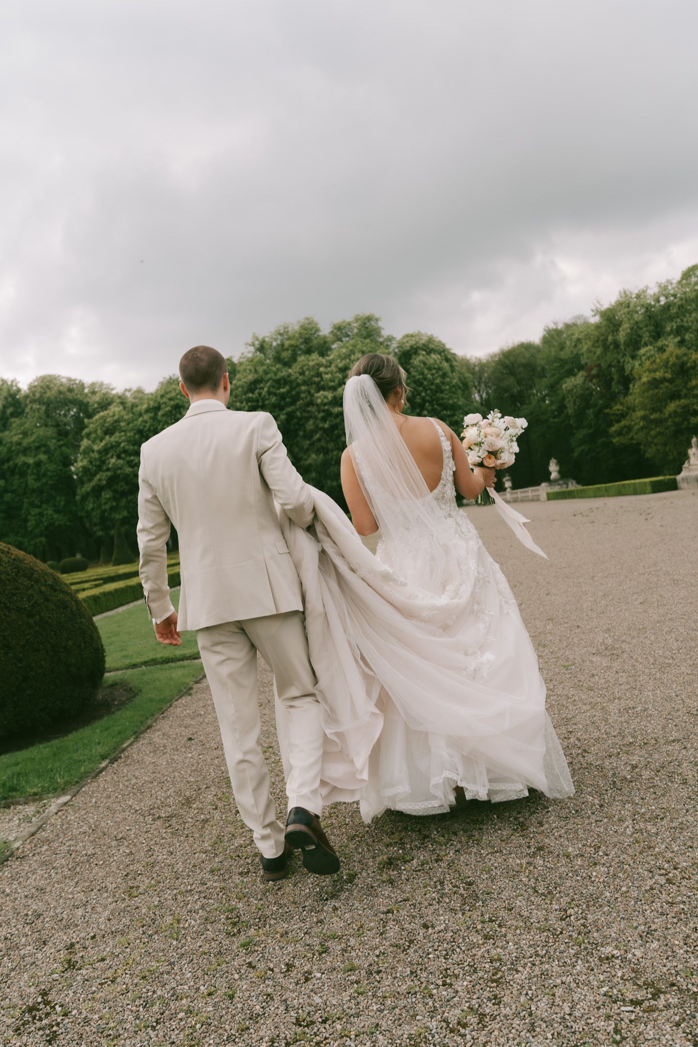 A wedding couple walks hand in hand across a gravel path surrounded by formal gardens in Germany.