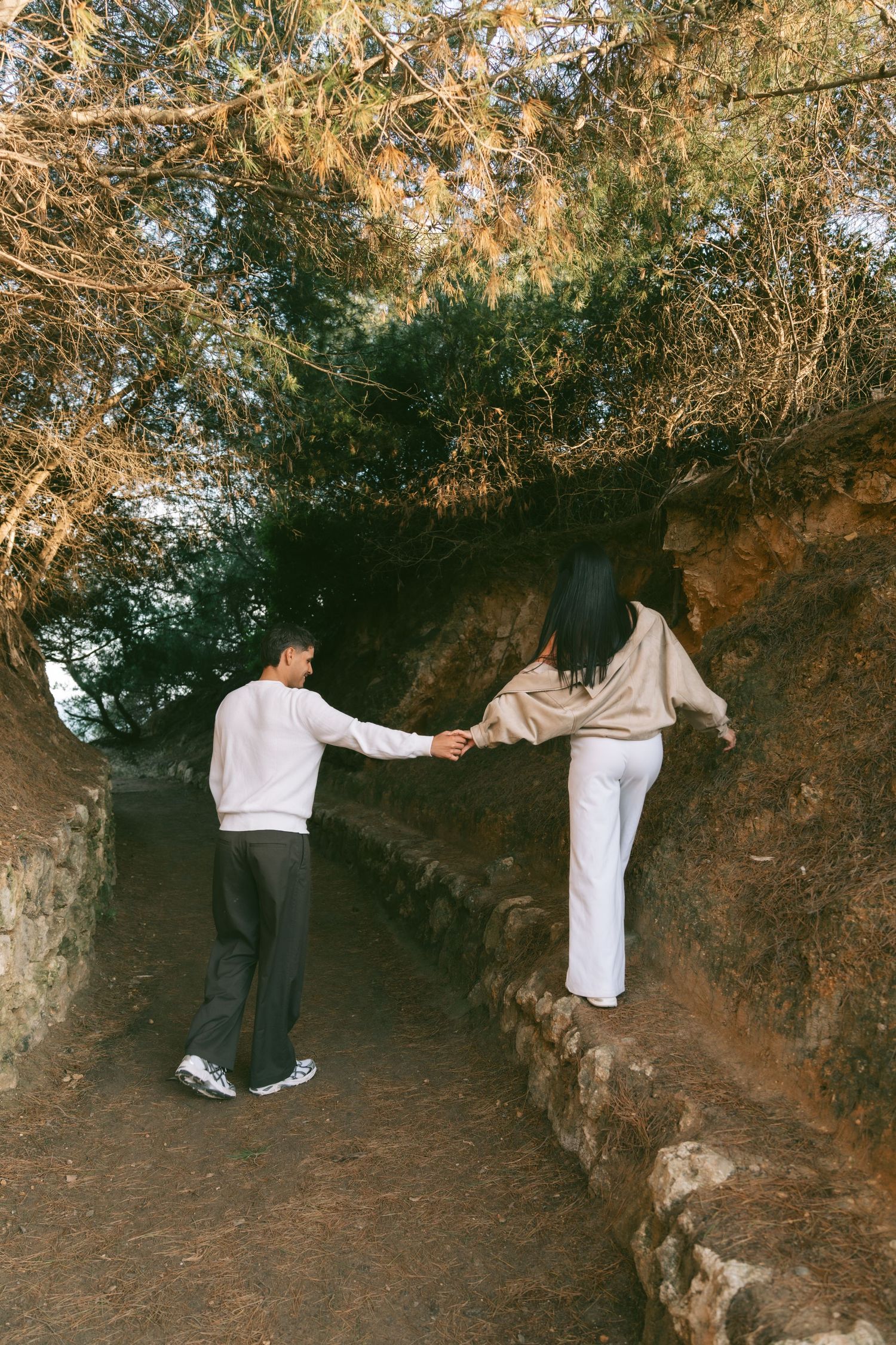 Couple holding hands on a rustic stone path under trees, man guiding woman up a natural trail.