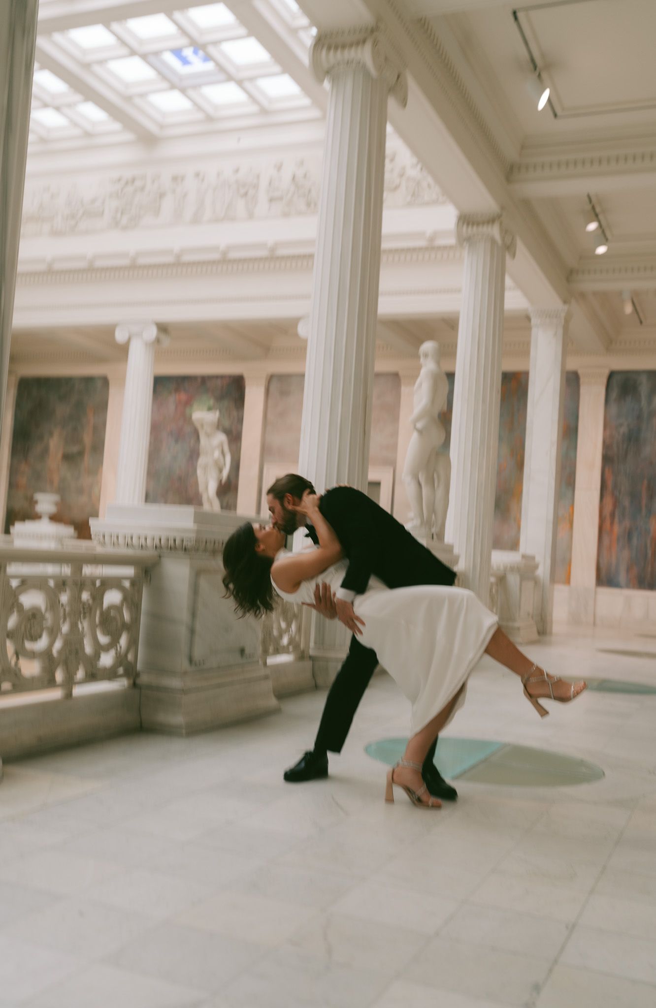 People perform artistic movements in a grand white marble hall with columns and skylight.