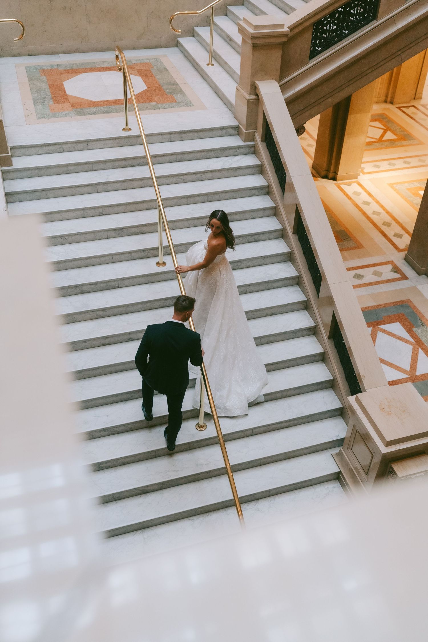 A bride and groom descend an elegant marble staircase in a grand building interior.