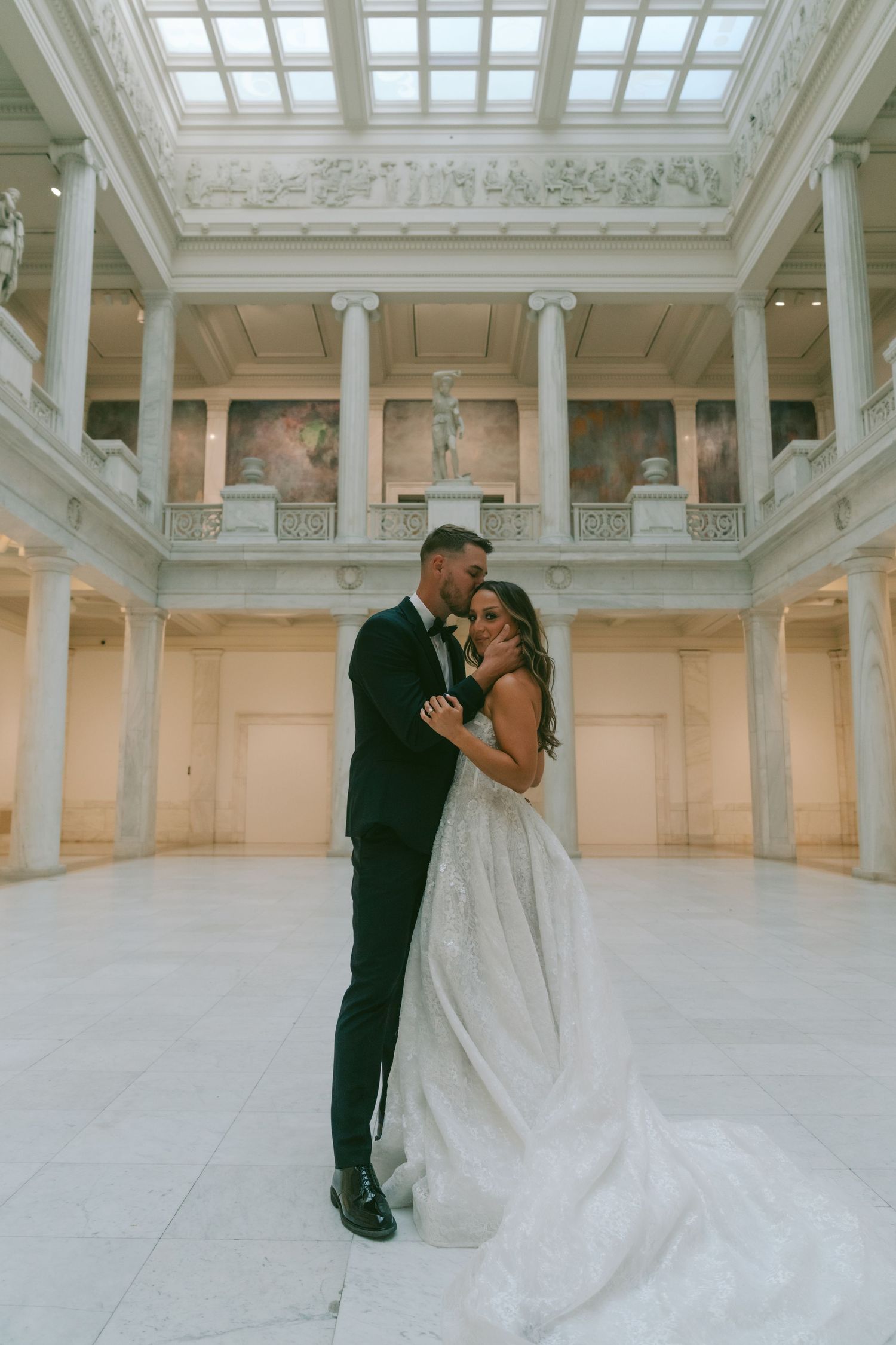 A romantic wedding couple shares intimate moments in an elegant white marble room with skylight windows.