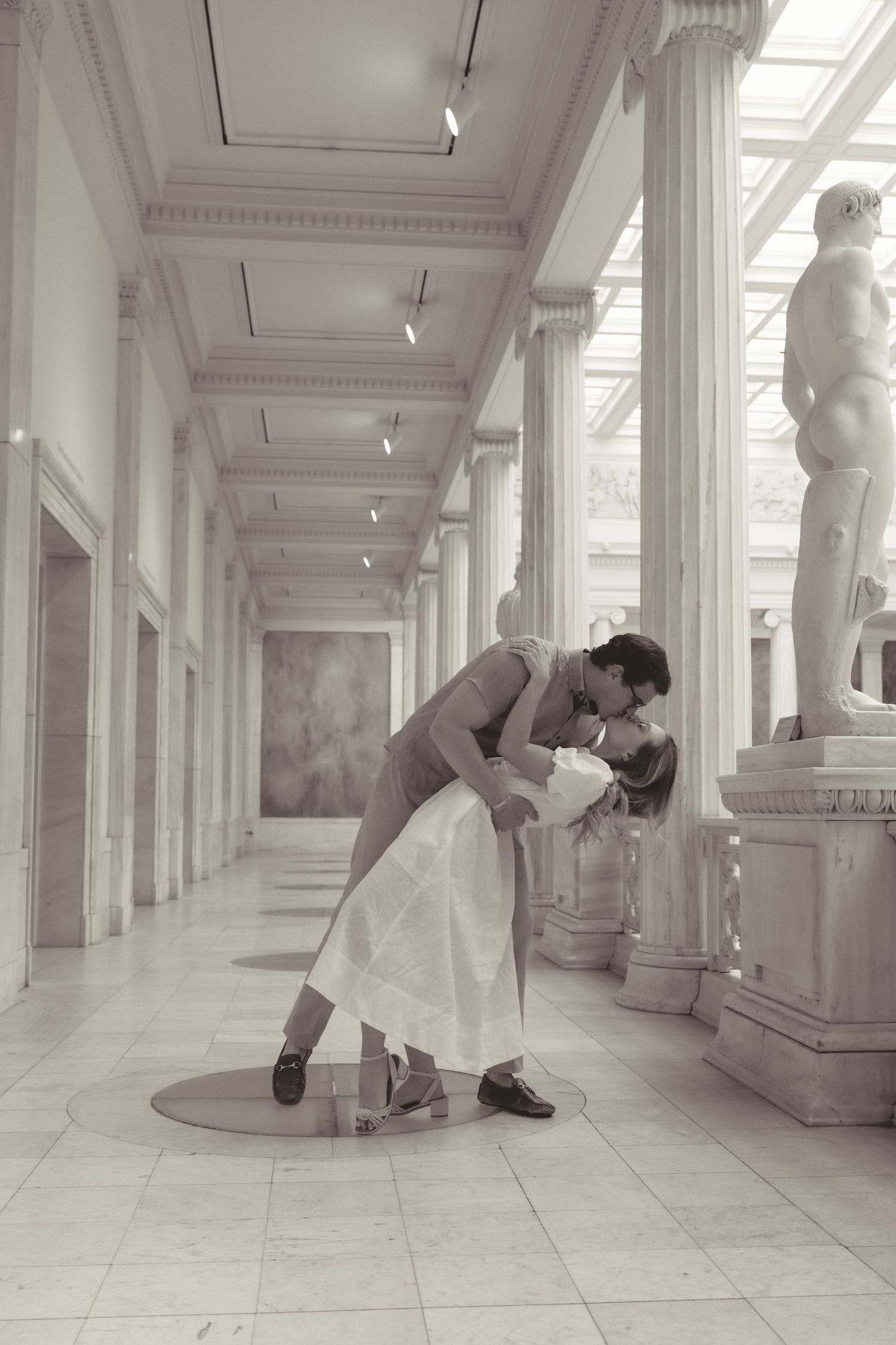A romantic dance pose captured in black and white in an elegant hallway with classical columns and marble statues.