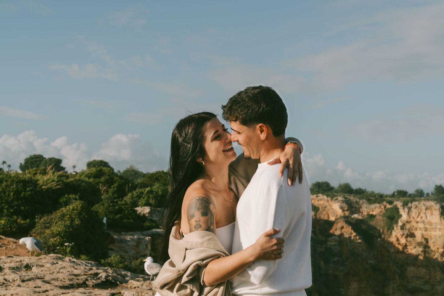 Happy couple embracing and laughing together on a rocky cliff with a scenic sky backdrop.