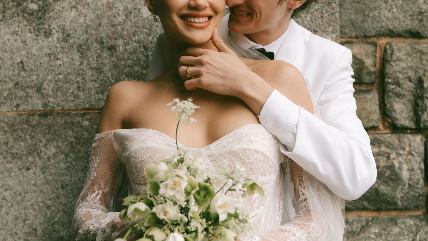 A smiling bride in a lace off-shoulder gown holds a white floral bouquet beside a groom in a white suit.