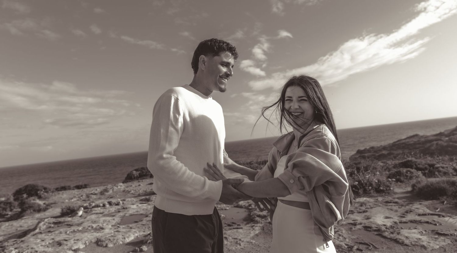 Black and white photo of a happy couple holding hands outdoors on rocky terrain with ocean views.