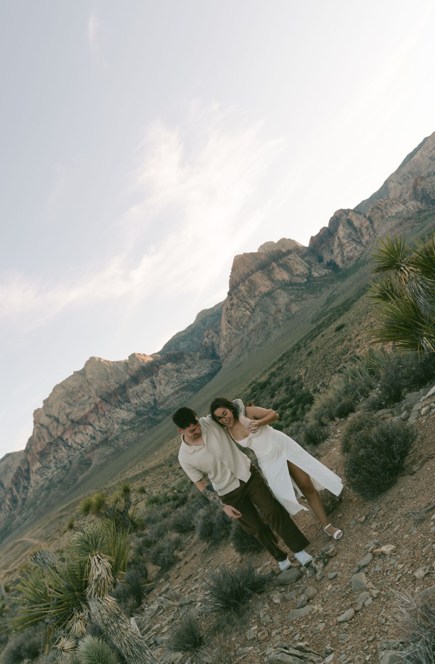 A couple in white outfits embrace on a rocky mountainside at sunset with dramatic cliffs in the background in Las Vegas, NV.