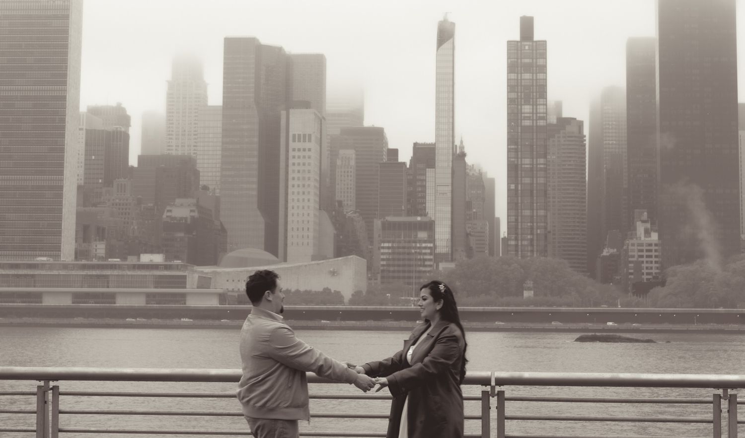Black and white photo of couple on waterfront with Manhattan skyline in background.