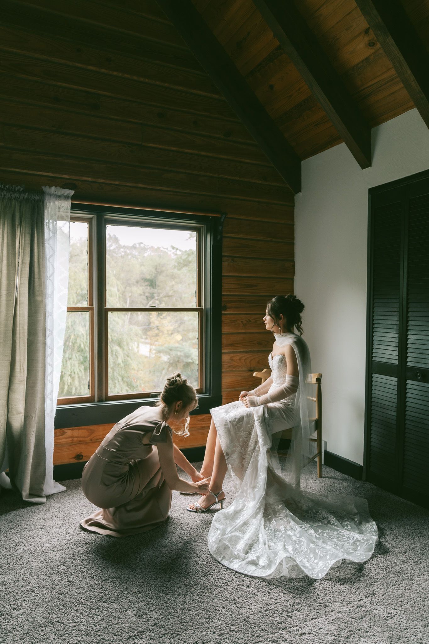 Two people sit by a window in a rustic cabin room with wooden beams and white curtains.
