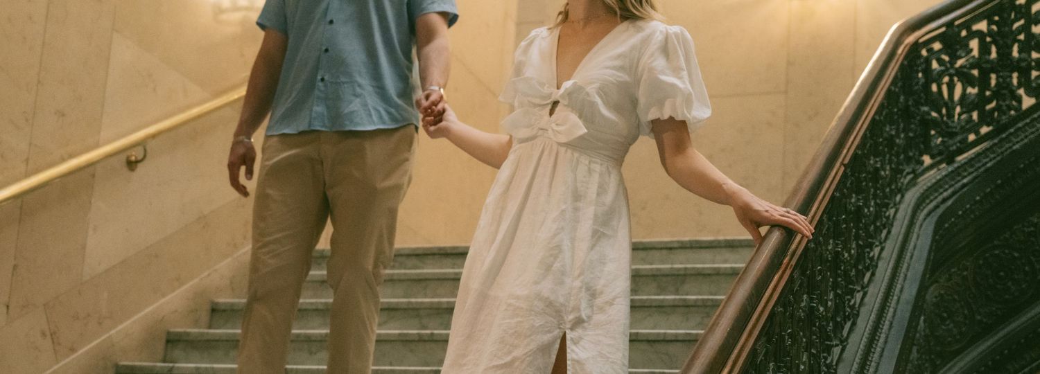A couple walks down an elegant staircase with ornate railings in vintage-inspired clothing.