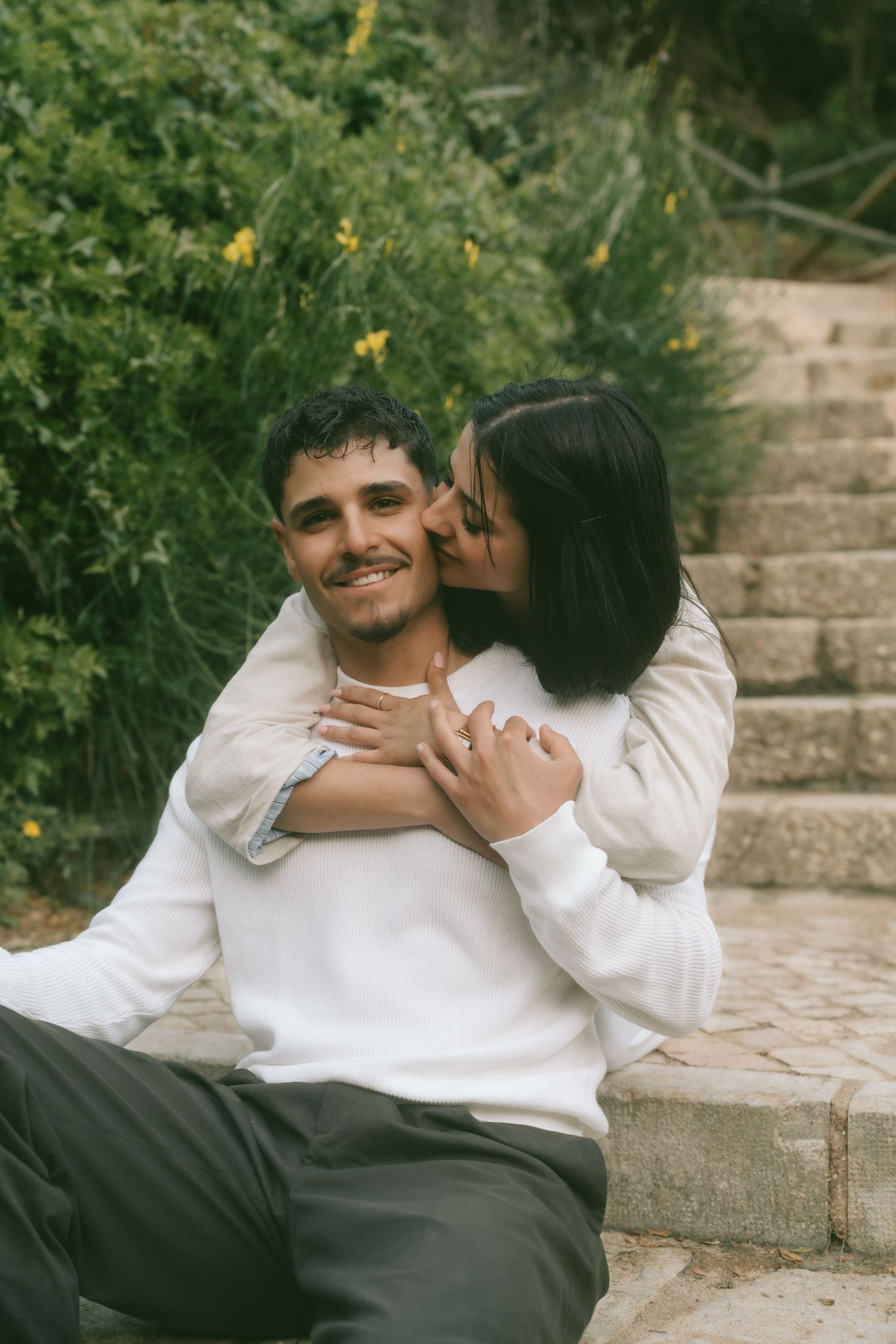 Happy couple embracing on stone steps surrounded by lush greenery, woman kissing mans cheek while he smiles.